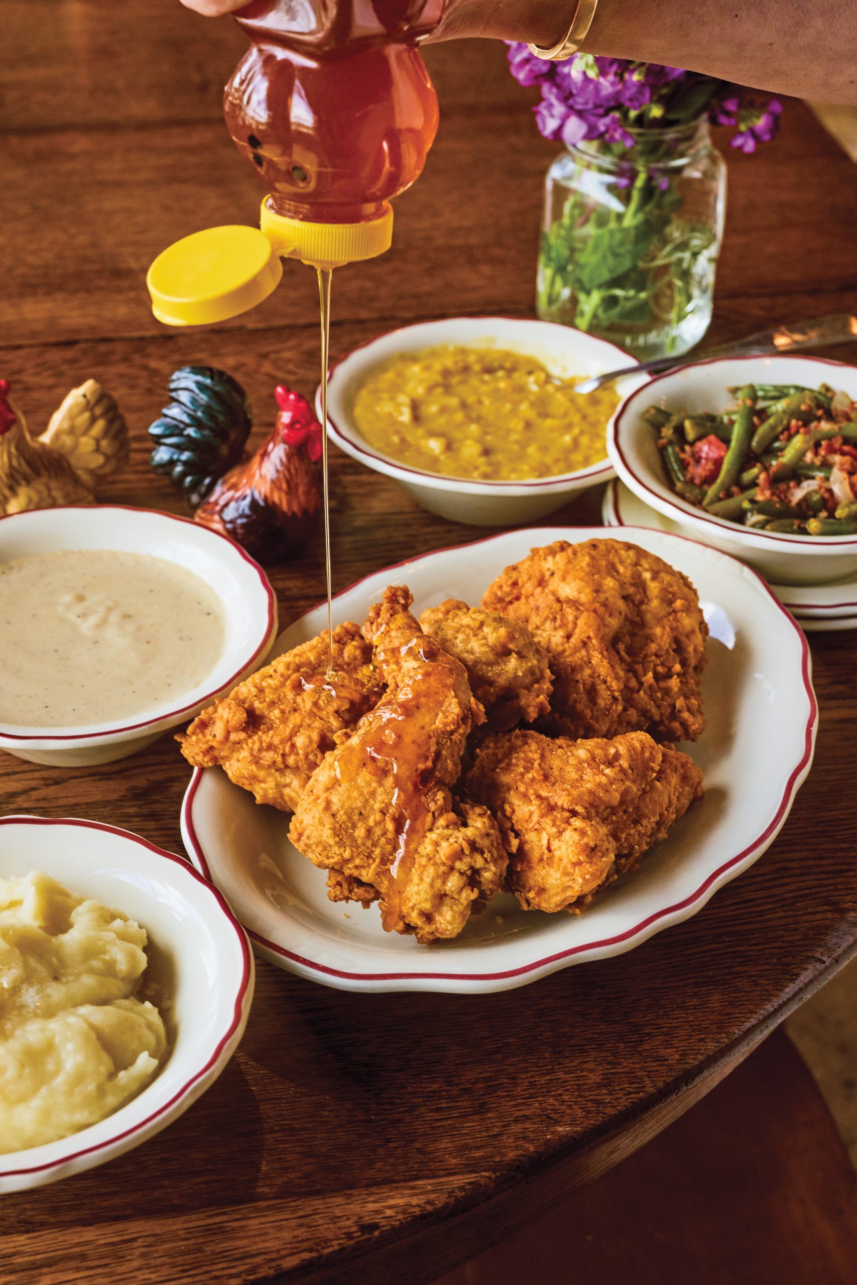 A bear-shaped bottle of honey is drizzled over a plate of fried chicken, surrounded by a spread of mashed potatoes, gravy, corn, and green beans. A jar of lilacs is also placed on the wooden table everything sits on.