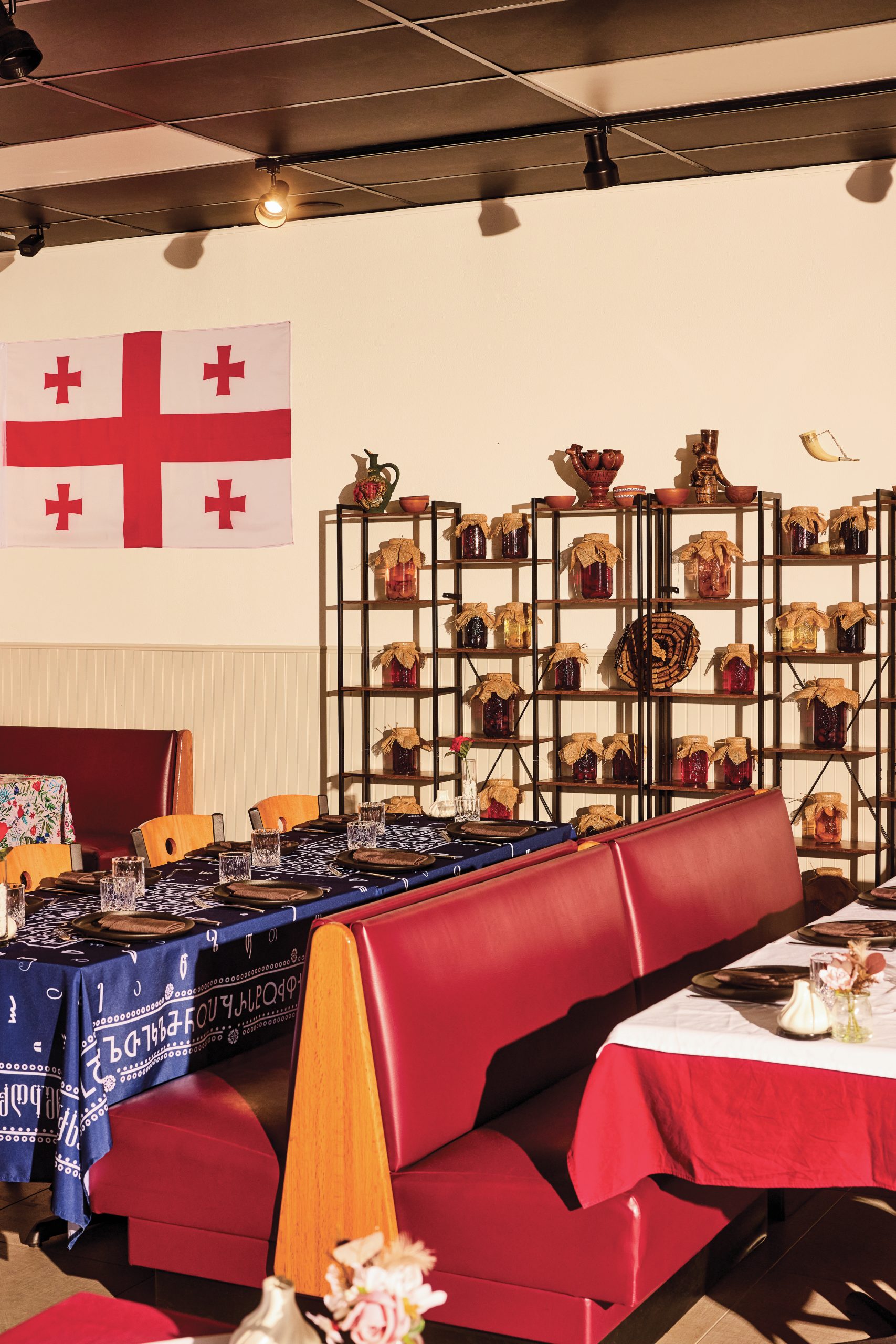 The interior of a Georgian restaurant, featuring red diner booth chairs and different table spreads of blue and white. Shelves of ingredients are seen in the back, on the right of the flag of Georgia, in front of white walls.