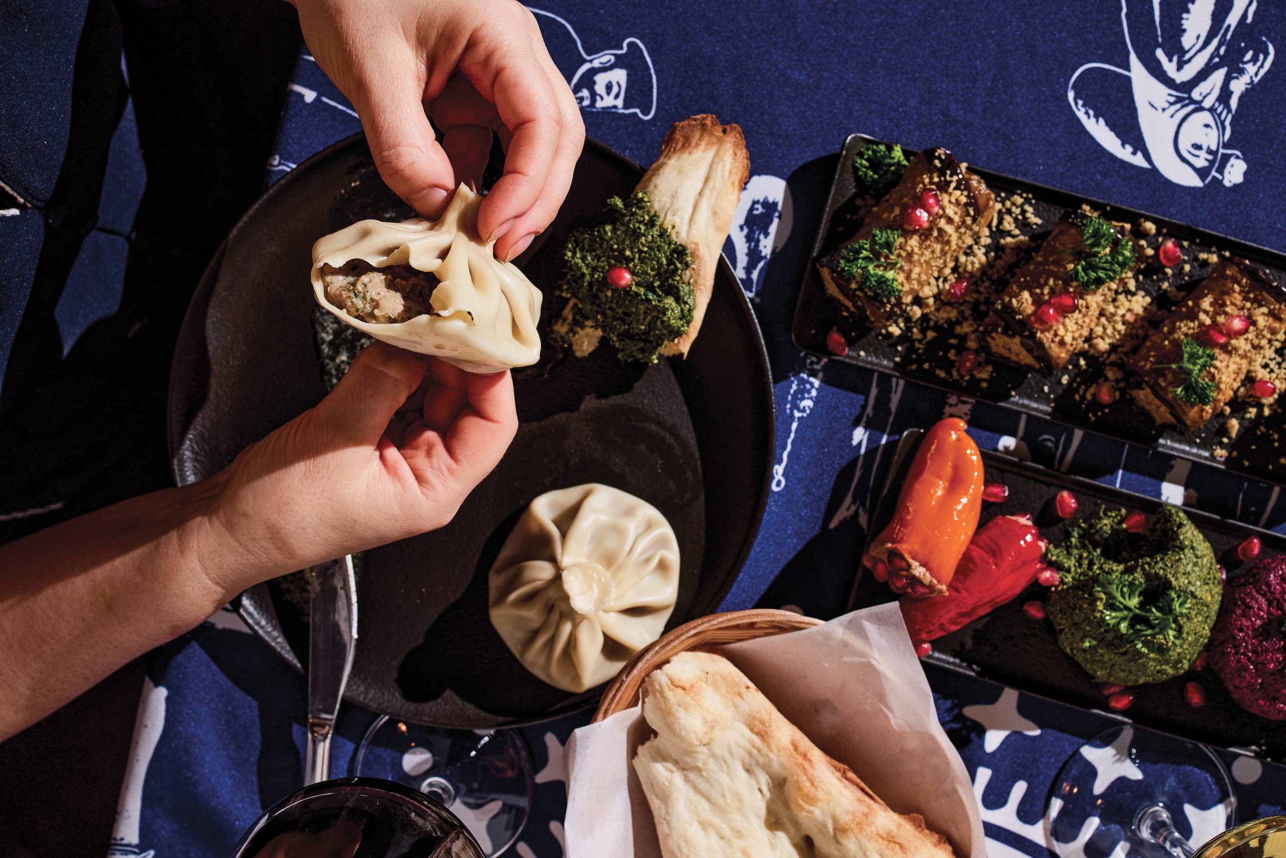 A vibrant spread of Georgian food, including dumplings, being plated by hand on a table with a dark-blue spread.