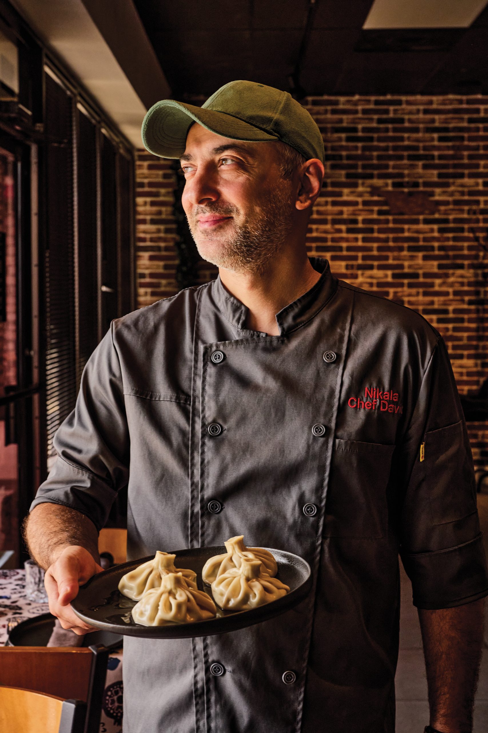 A smiling adult Georgian man wearing a dark gray chef's jacket and olive-green cap looking to the left while holding a plate of dumplings.