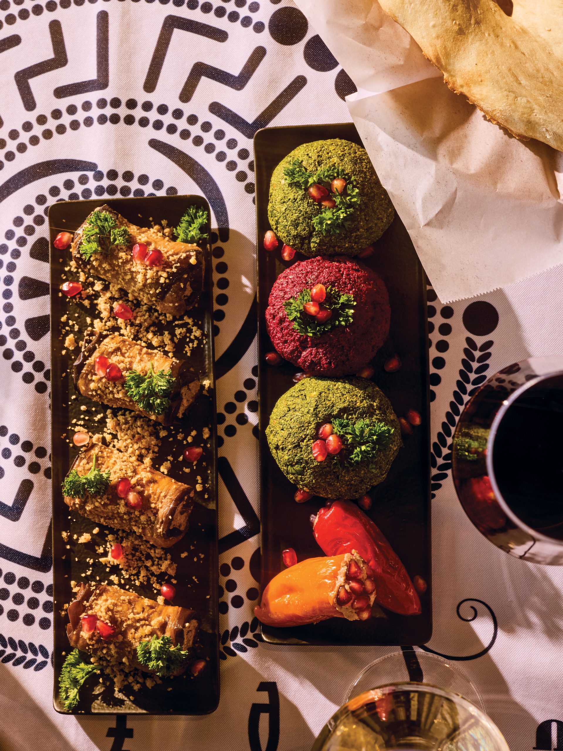 Two black rectangular plates, one with four eggplant rolls and the other with four colorful pkhali, placed on a table with a white-and-black patterned spread.