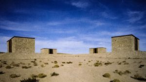 A wide-angle photograph of James Magee's The Hill, a large-scale art installation featuring four stone buildings in a cross shape on desert land against a bright blue sky.