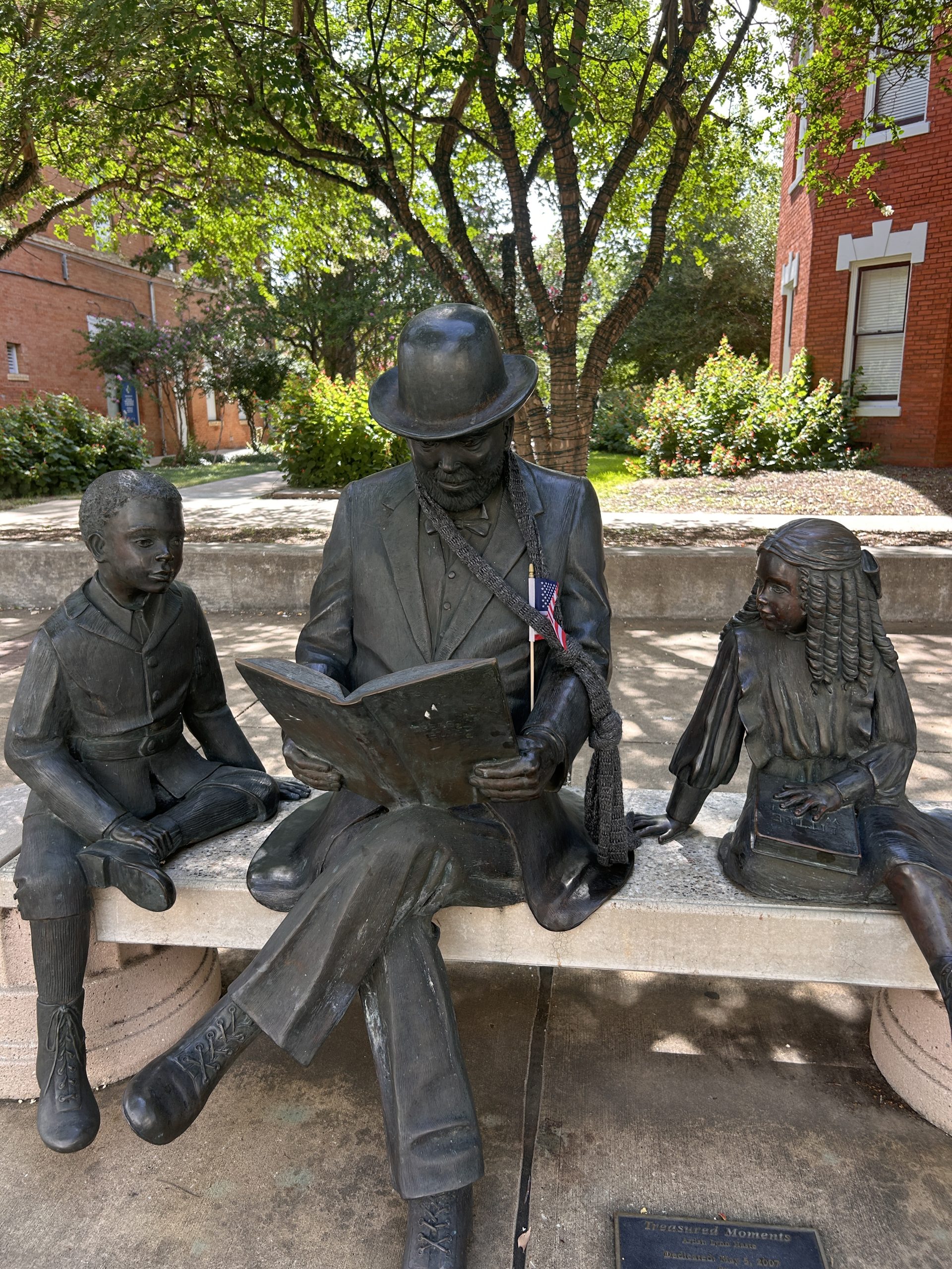 Bronze sculpture of man wearing bowler hat and three-piece suit reading book and sitting between two children on bench.