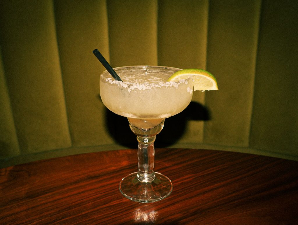 A margarita on the rocks with salt on the rim of the glass and a lime wedge with a black straw sticking out on a wooden table