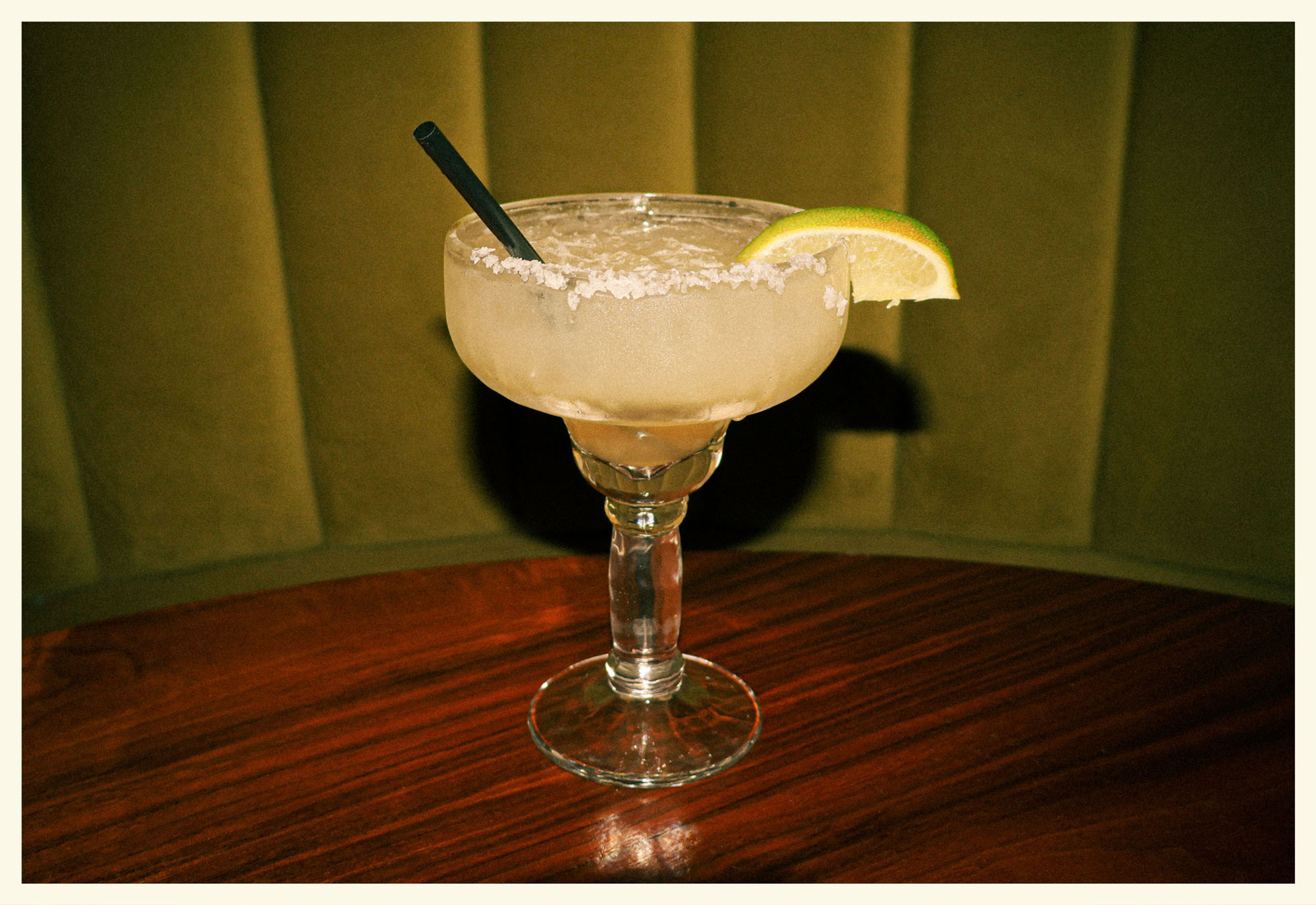 A margarita on the rocks with salt on the rim of the glass and a lime wedge with a black straw sticking out on a wooden table