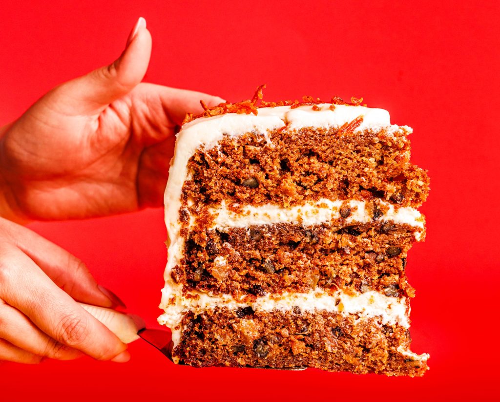 A woman's hands hold a large three-layer slice of carrot cake in front of a bright red backdrop