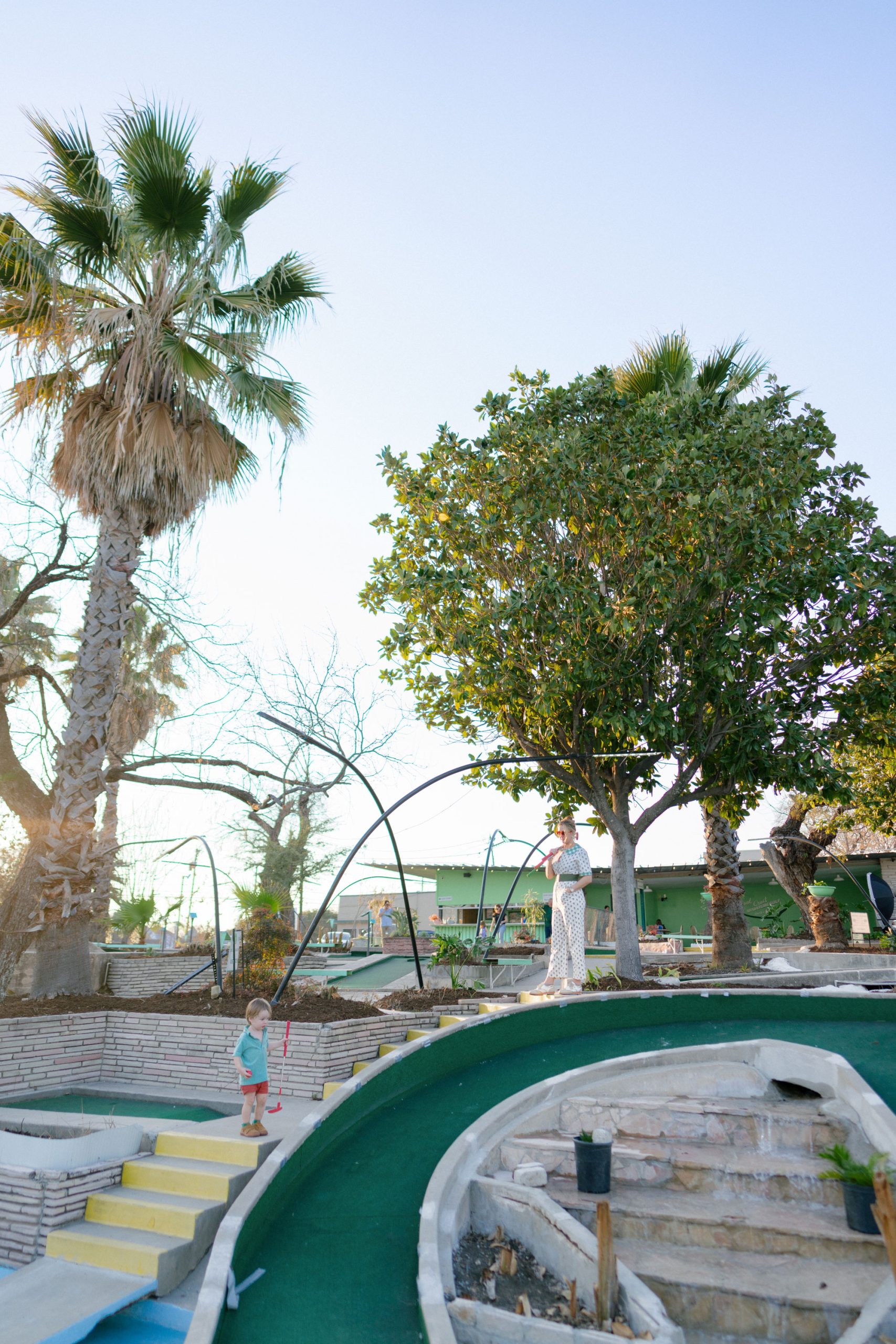 A woman and toddler stand on concrete stairs with a part of a mini golf course in front of them and trees behind them.