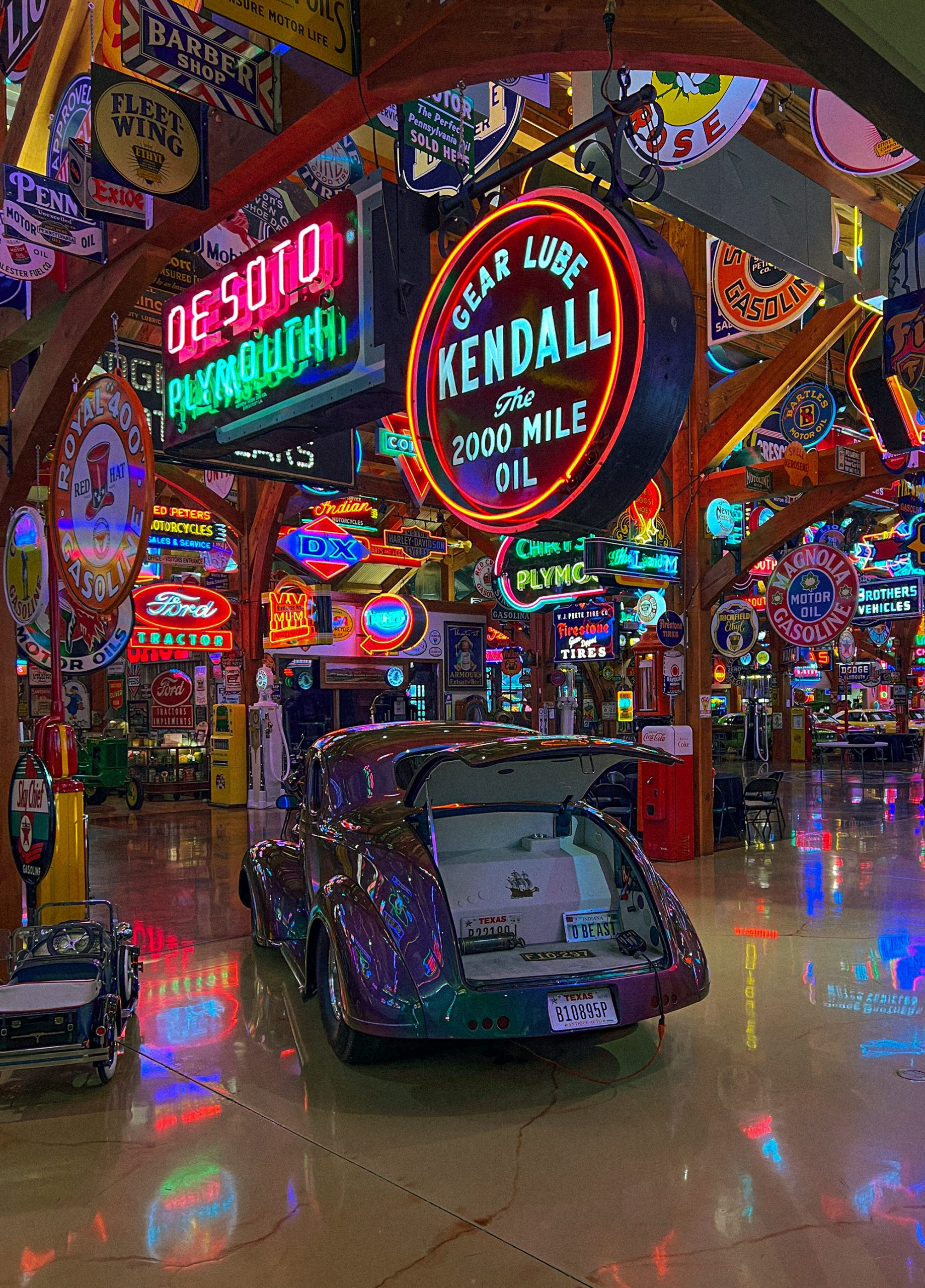A vintage car with its trunk open sits inside a building where the roof and walls are covered by vintage neon and porcelain signs.