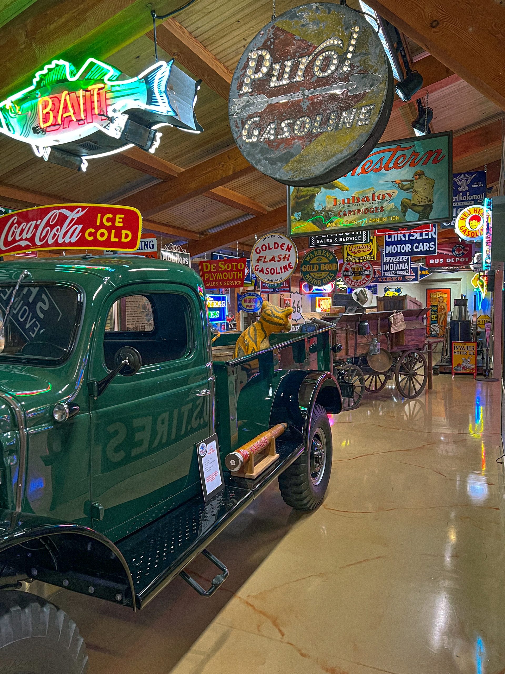 A vintage green truck sits inside a building filled with vintage signs, including a neon fish sign that says "Bait" and a circle sign that says "Purol Gasoline."