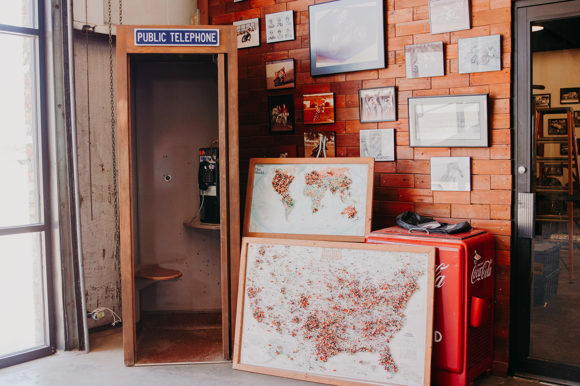 A framed map of the the world and a framed map of the United States filled with pins marking locations. The maps are up against a wall full of framed photos and beside an old public telephone booth