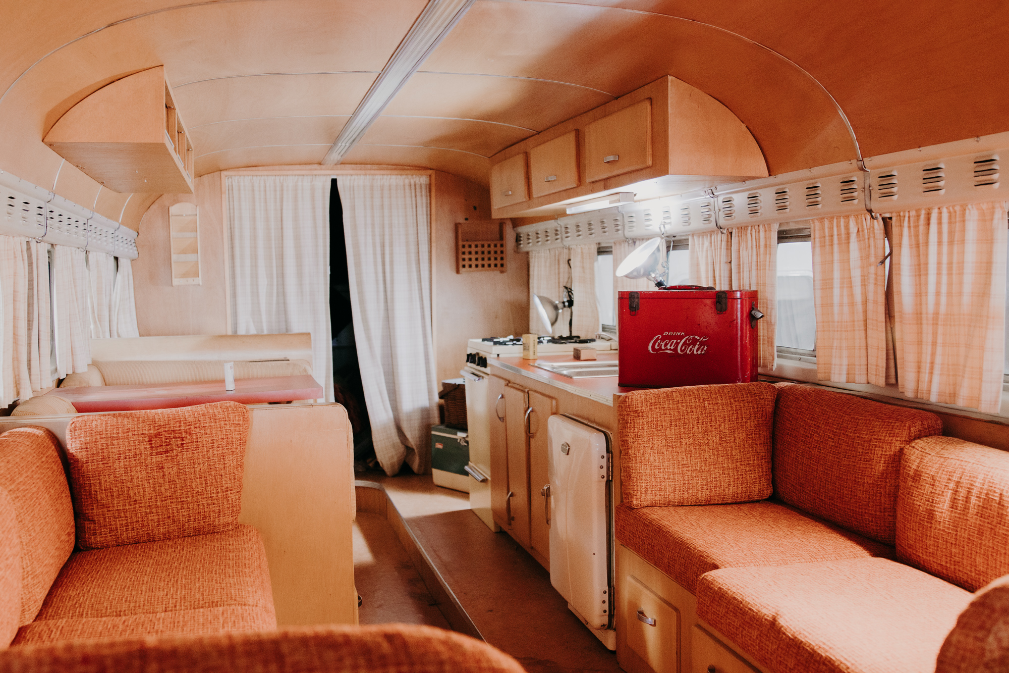 The inside of a retro RV furnished with wood paneling and orange cushions. A Coca Cola cooler sits on the counter