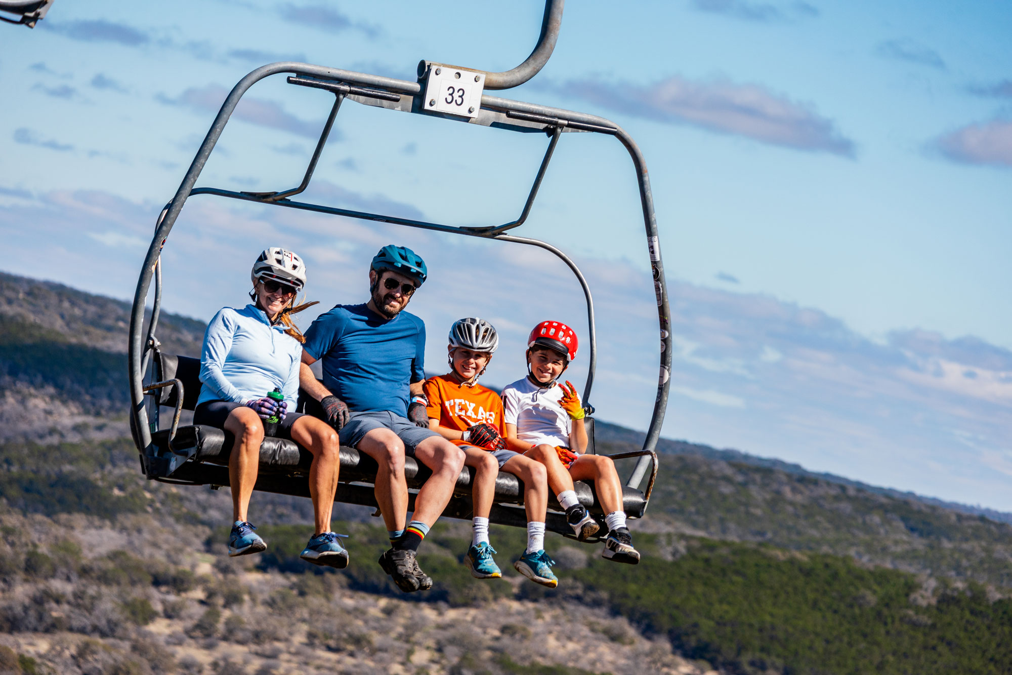 A family of four sits on a chairlift with the sky and ground behind and beneath them.