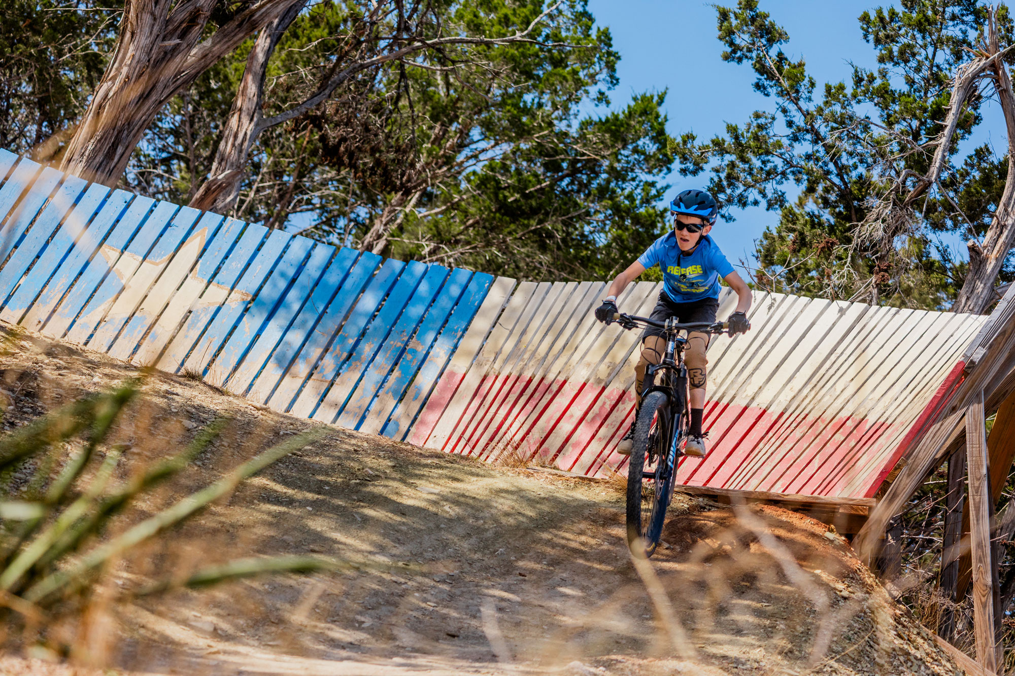 A little boy in a blue shirt and helmet rides off of a wooden wall that is painted to look like the Texas flag.