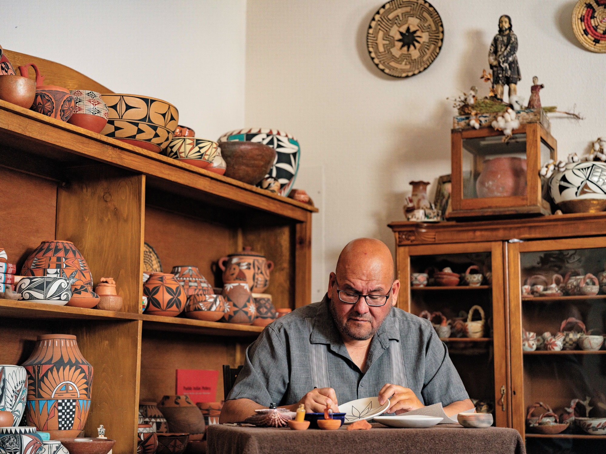 A man works on a piece of pottery inside a studio surrounded by more pieces of pottery.