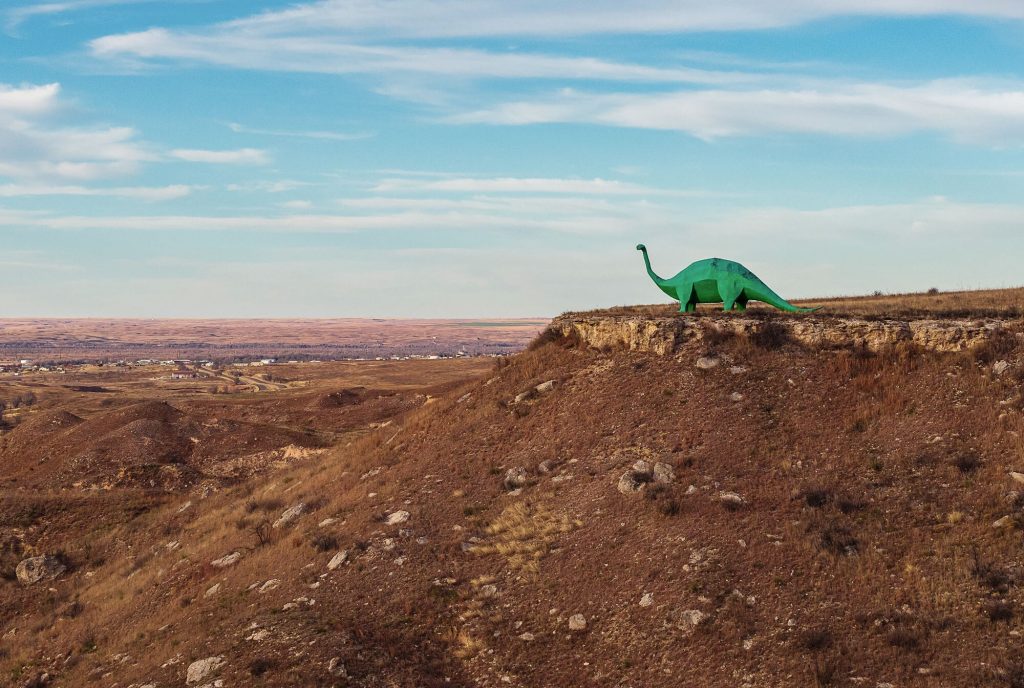 A green dinosaur statue peers over a brown rocky cliff