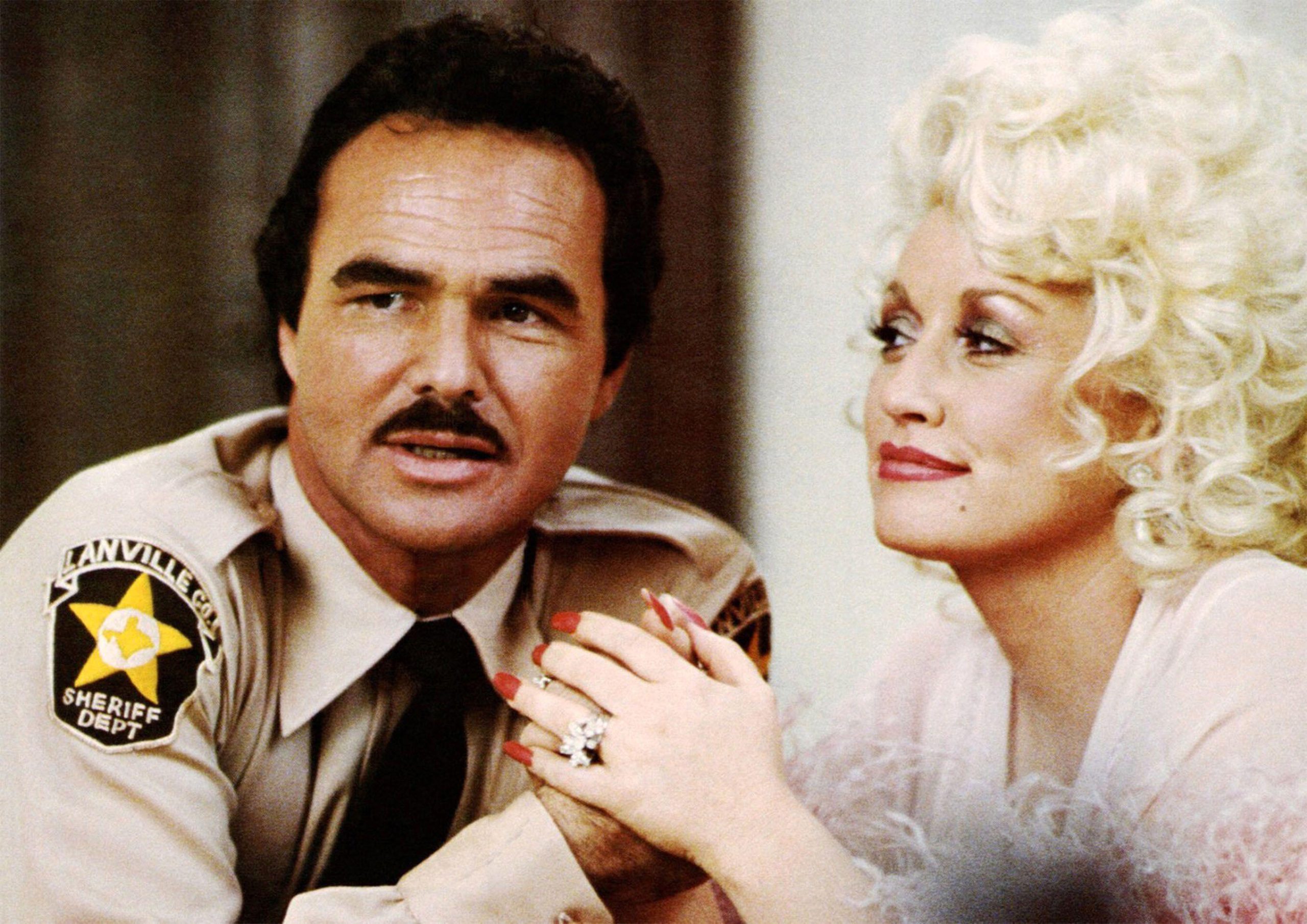 A man in a tan sheriff's uniform holding hands with a blonde woman wearing a sparkling ring
