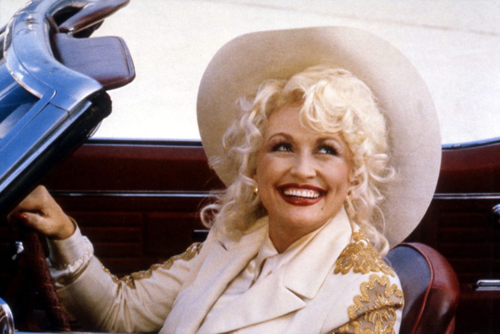 A blonde woman wearing bright red lipstick, a cowboy hat, and cream-colored Western wear with gold flourishes smiles while driving a convertible