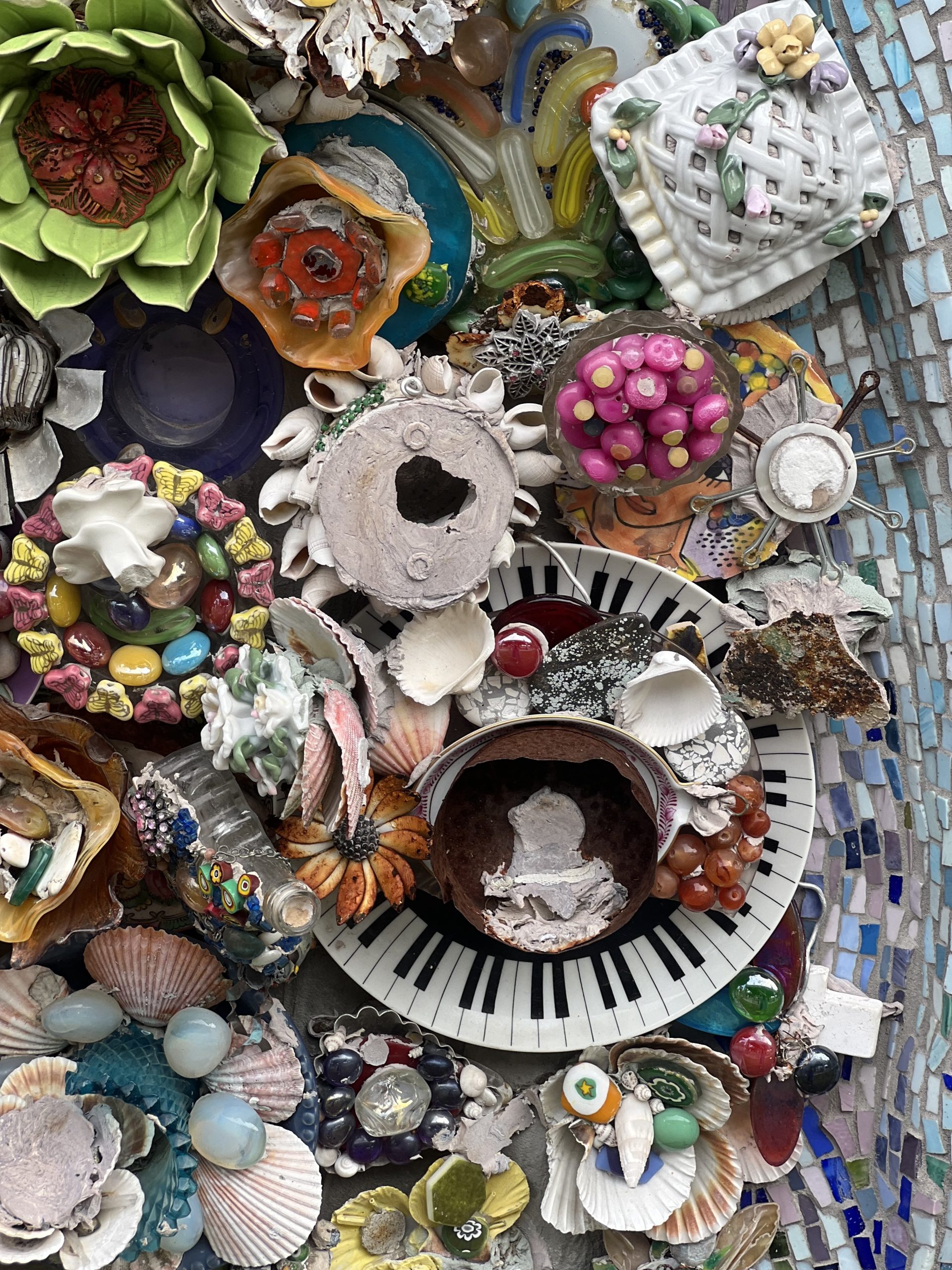 A colorful mosaic with colorful ceramic flowers, seashells of all kinds, painted rocks, baubles, a plate with a piano keys border, a bowl of ceramic radishes.