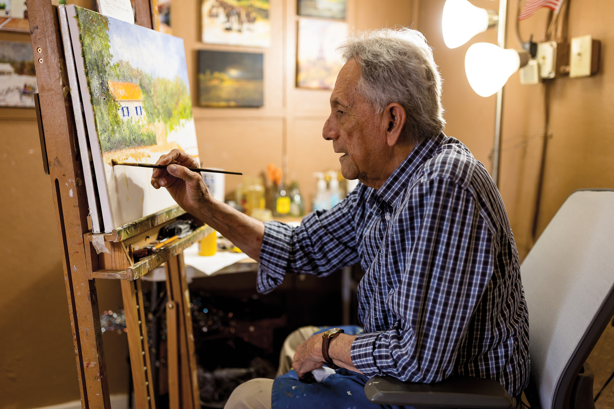 A man with white hair in a blue button-down shirt painting on a canvas that is resting on an easel