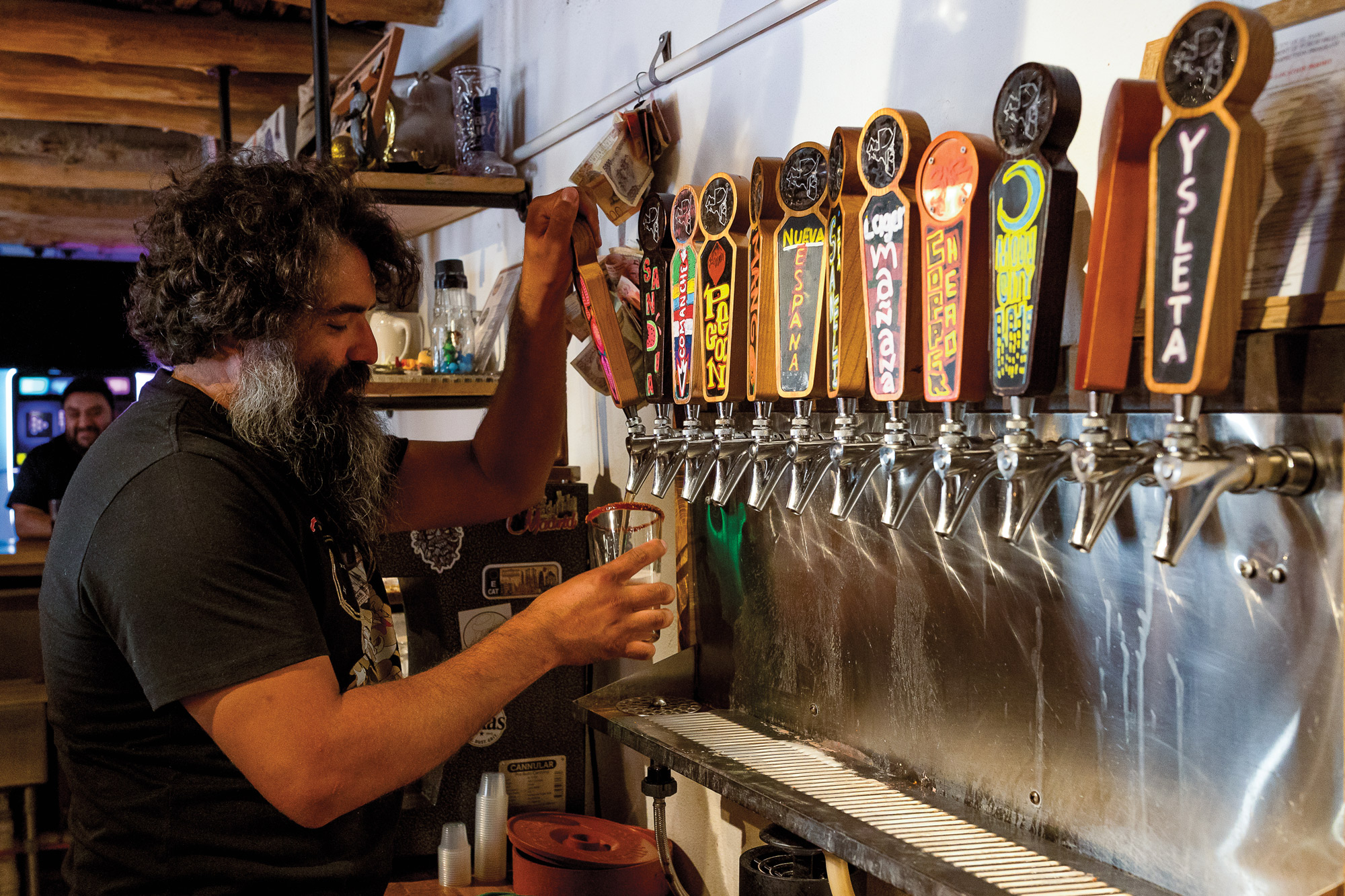 A bearded man pouring a beer from a tap among about 10 colorful taps behind a bar