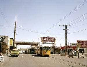 A yellow streetcar in the middle of a street that is lined with people and which has a red sign visible above that reads PARK HERE