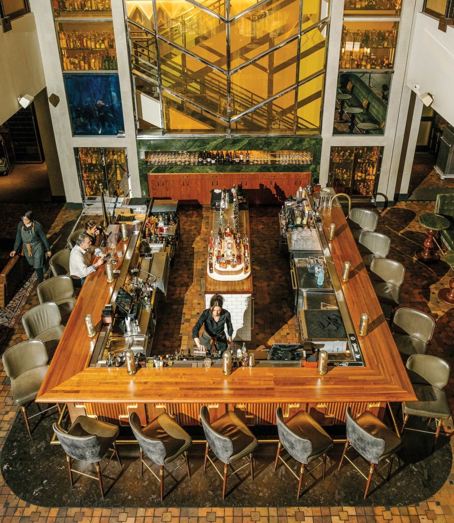 A woman in black mixes a cocktail behind a large wooden rectangular bar, with two customers sitting next to each other on one side and a staircase visible behind all