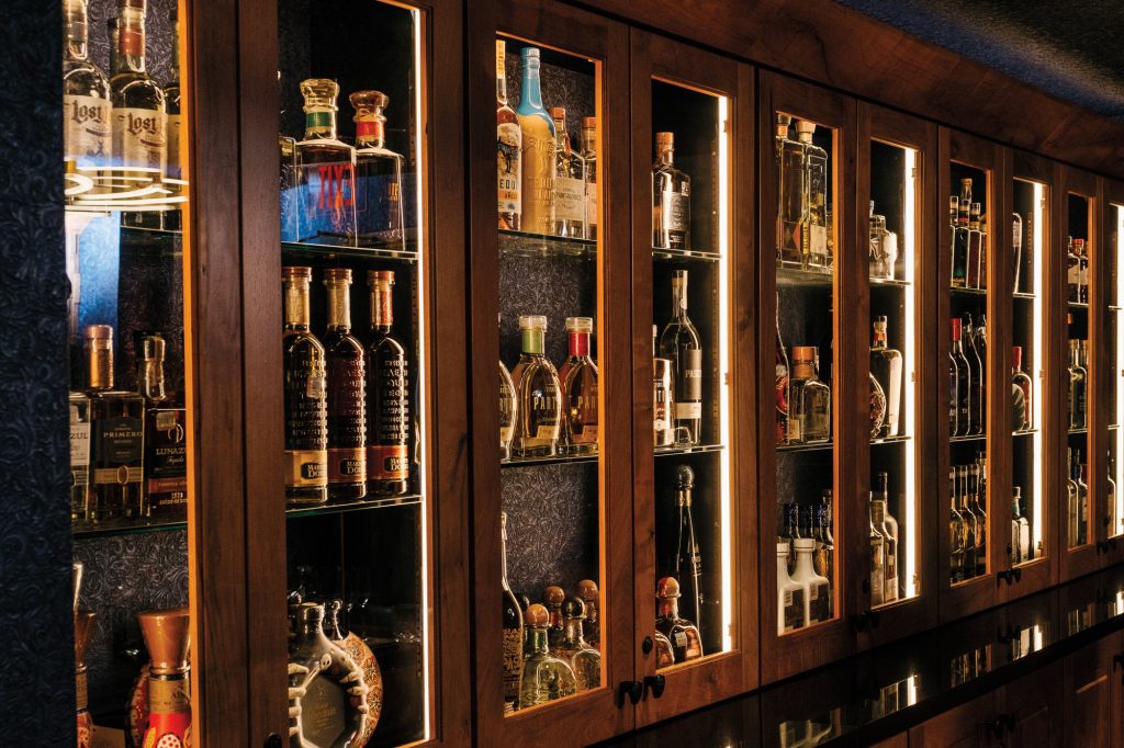 A wooden liquor cabinet filled with bottles of brown and clear liquor with three shelves in each