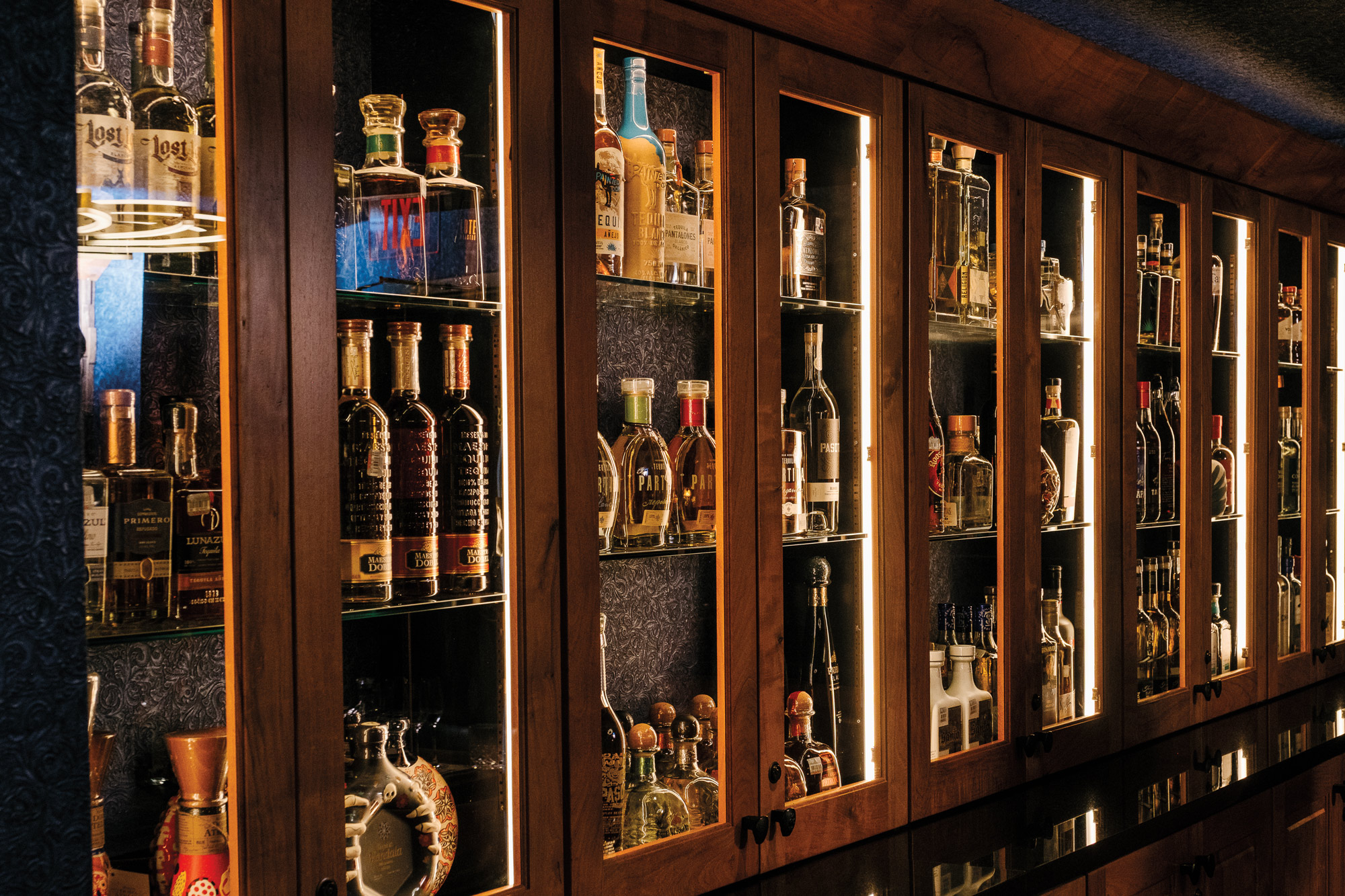 A wooden liquor cabinet filled with bottles of brown and clear liquor with three shelves in each