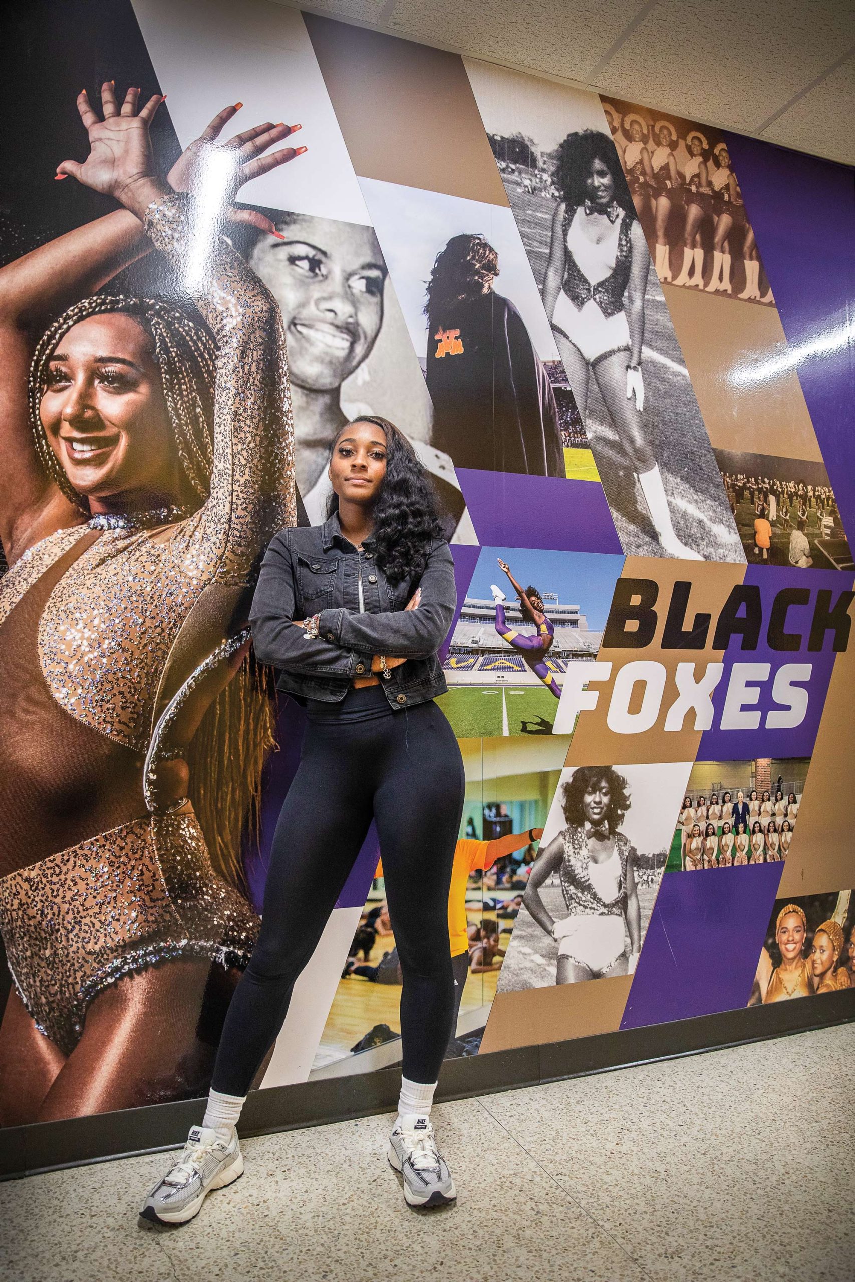 A woman in black leggings and a black denim jacket with her arms folded standing in front of a mural with the words Black Foxes visible