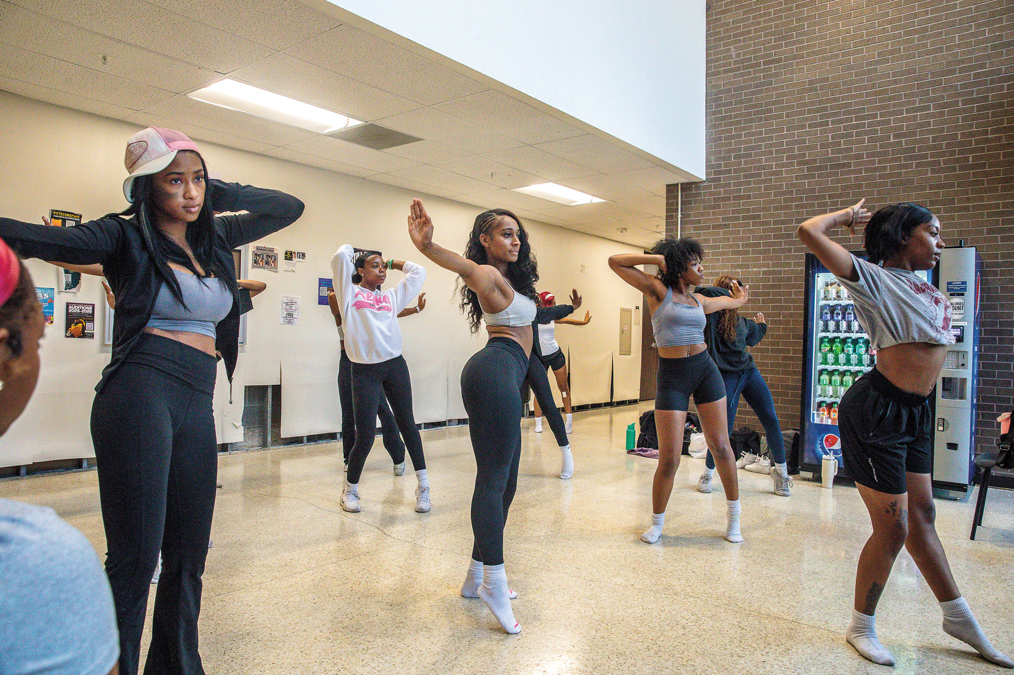 Women in various dance poses in a hallway in front of a soda vending machine