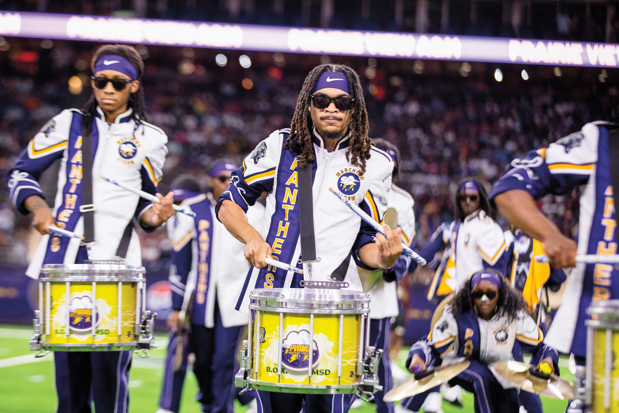 A marching band featuring drummers, in purple and white uniforms with the word Panthers visible