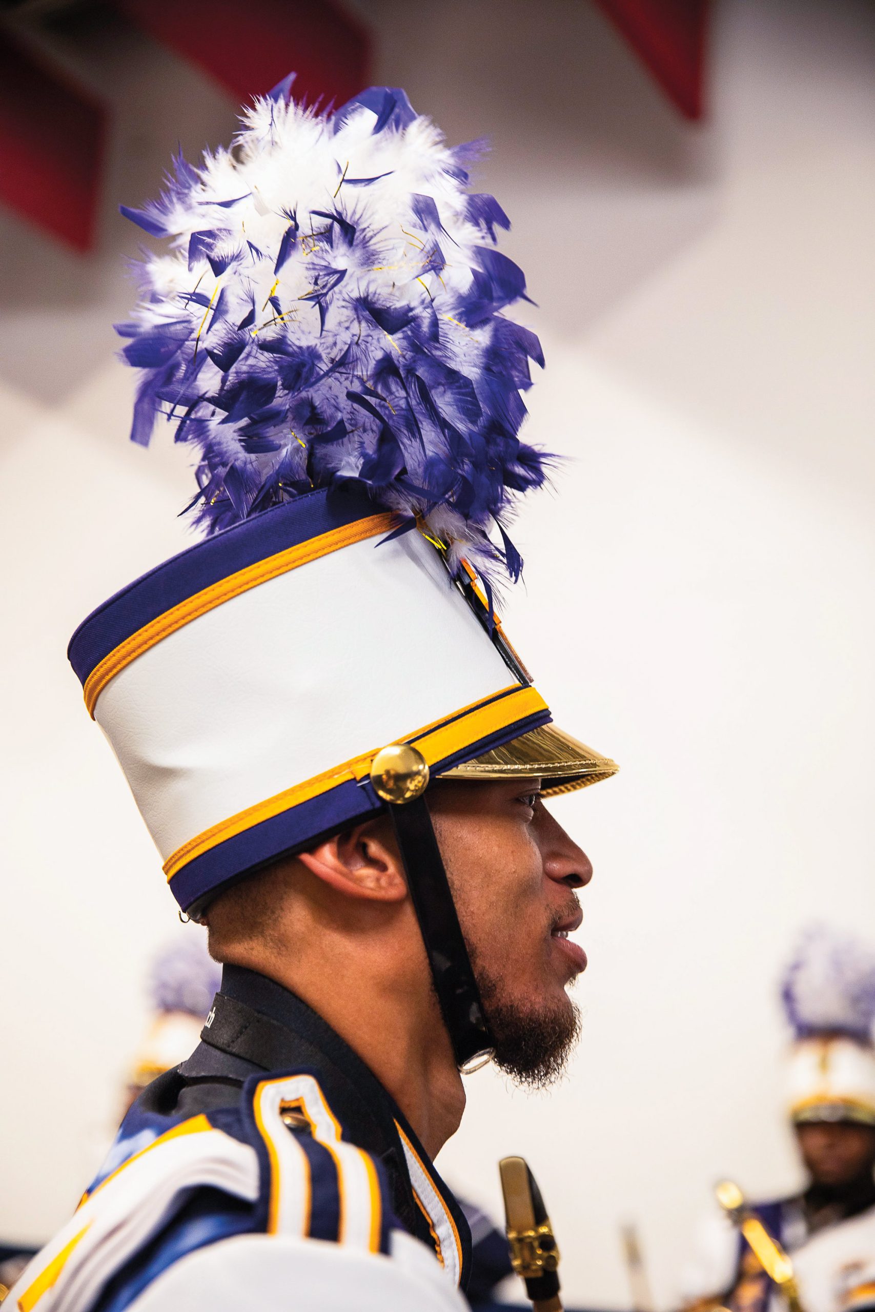 A marching band member wearing an ornate white, purple, and yellow cap