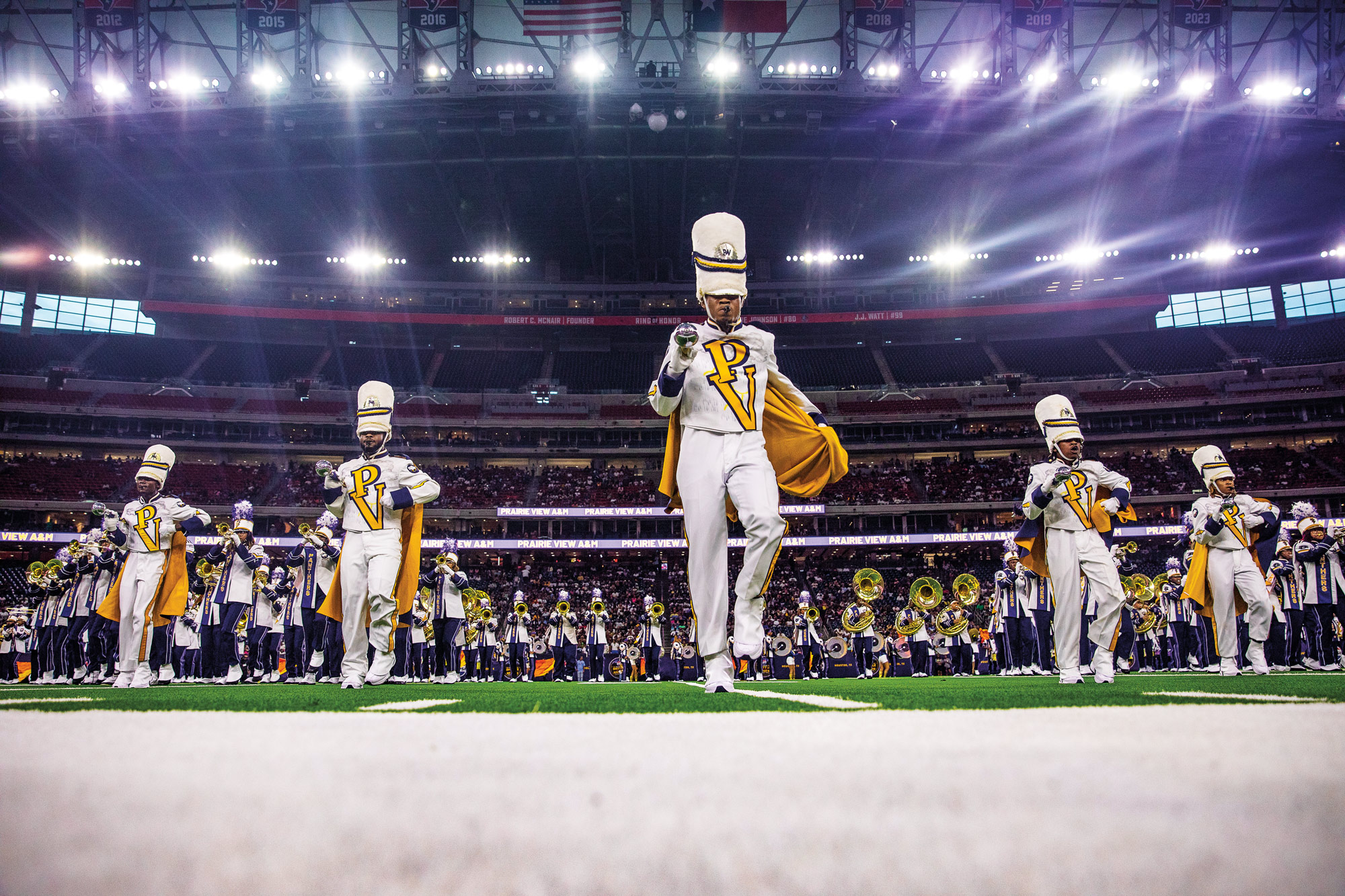 A marching band wearing white and purple uniforms on a football field in a large stadium