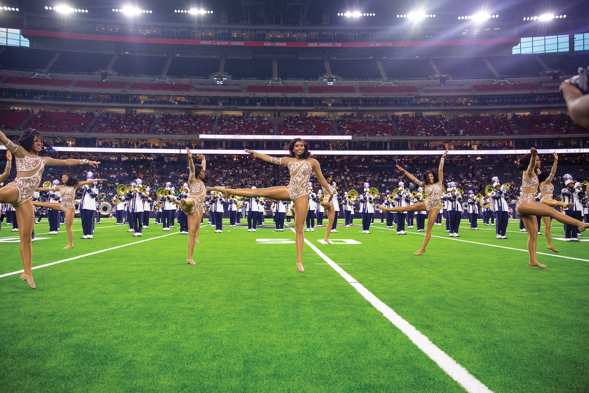 A dance team on a football field in front of a marching band inside a large stadium
