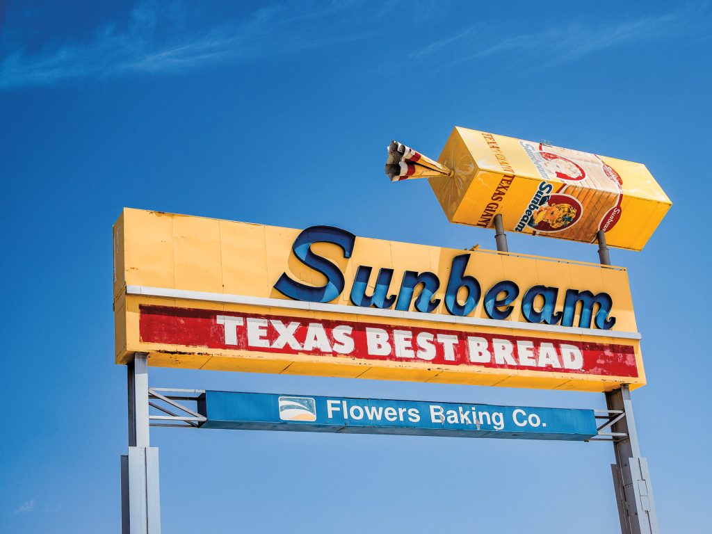 A yellow, red, blue, and white sign with a large display of a bag of bread, with the sign reading Sunbeam Texas Best Bread Flowers Baking Co.