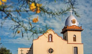 A light tan painted church with a silver belfry containing a silver cross atop it