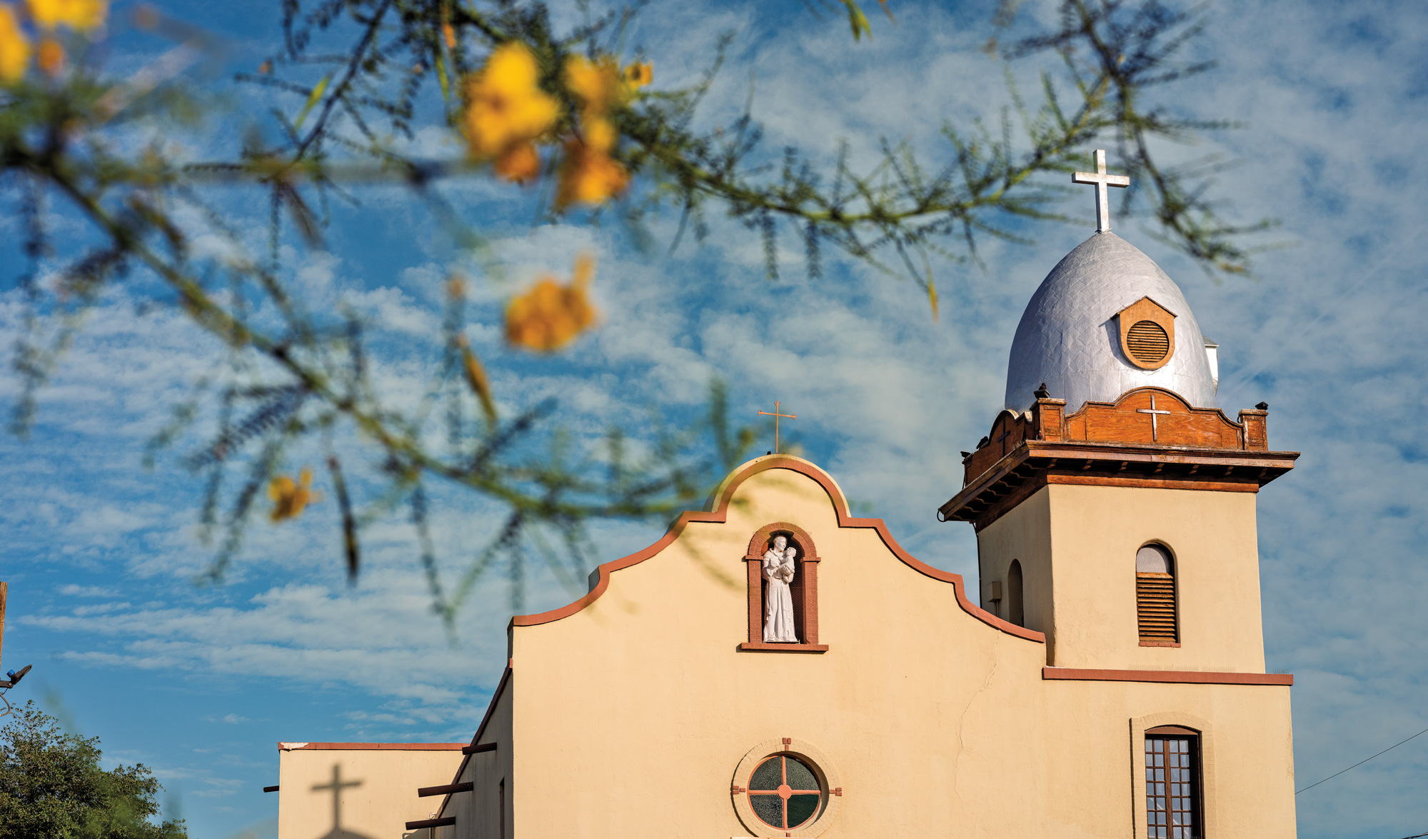 A light tan painted church with a silver belfry containing a silver cross atop it
