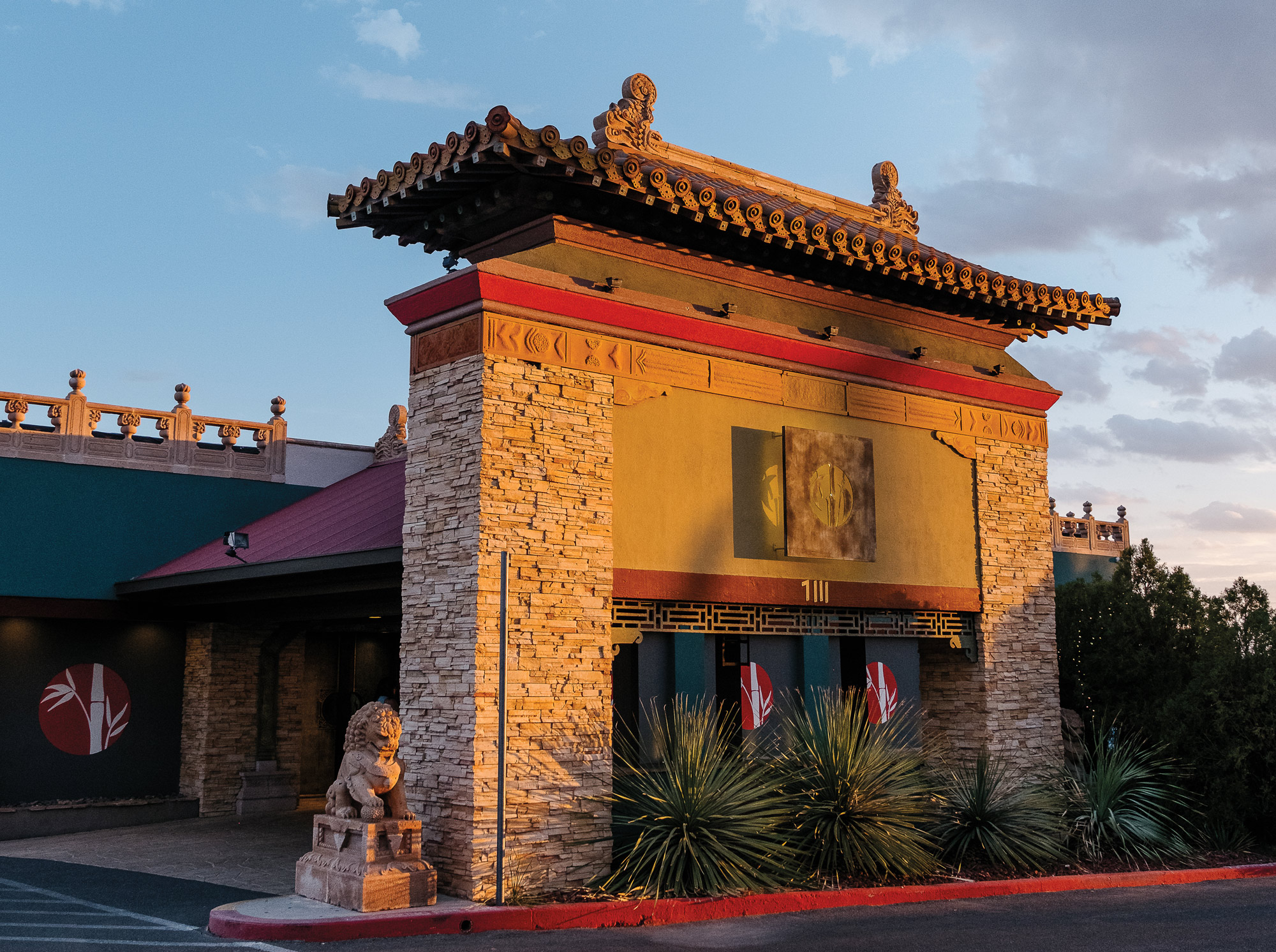 An exterior wall of a restaurant, featuring ornate stonework, a red roof, and red trim, with lush green plants in front of it
