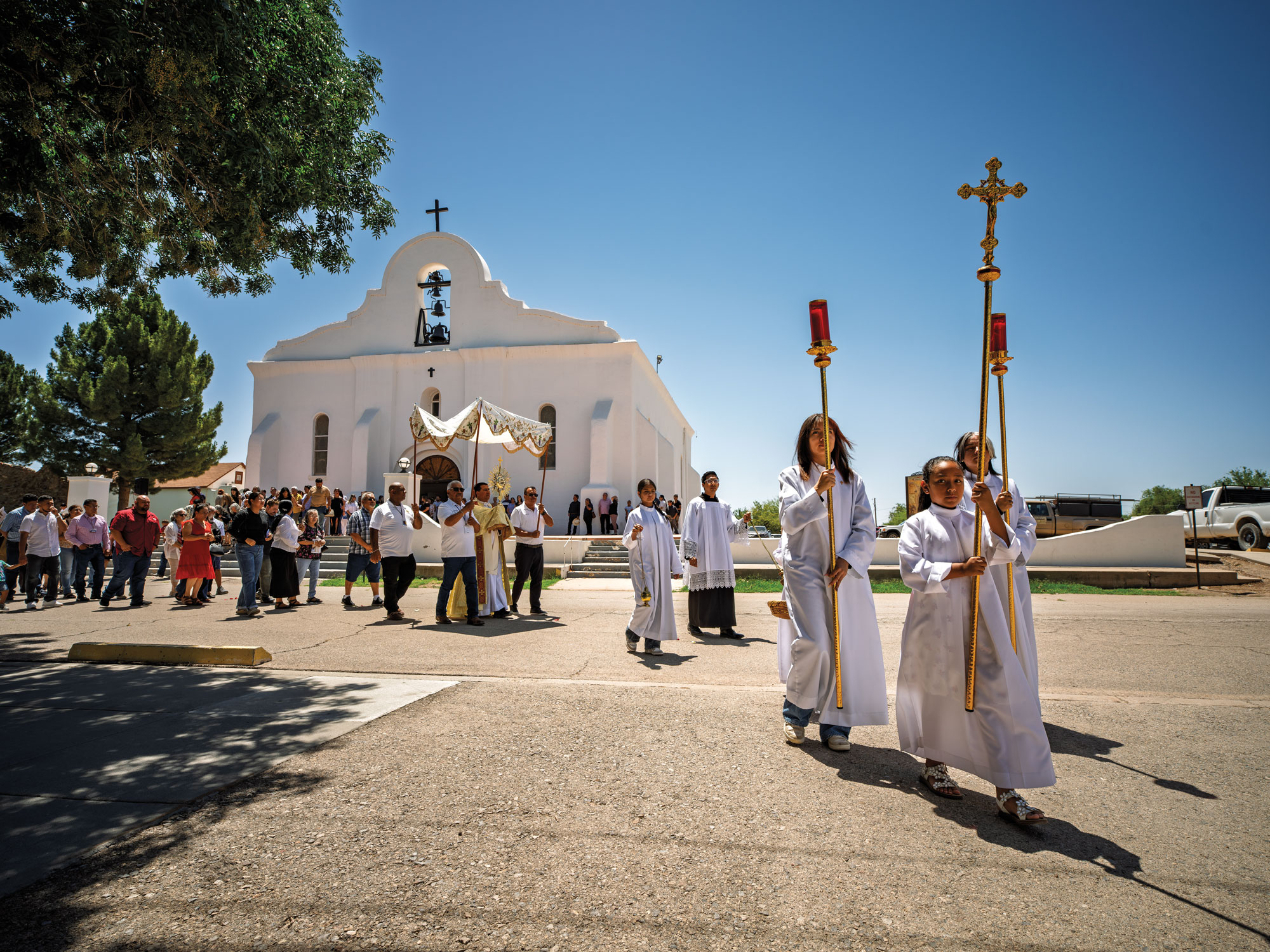 People dressed in white church robes holding a cross and candles lead a procession in front of a white chapel.