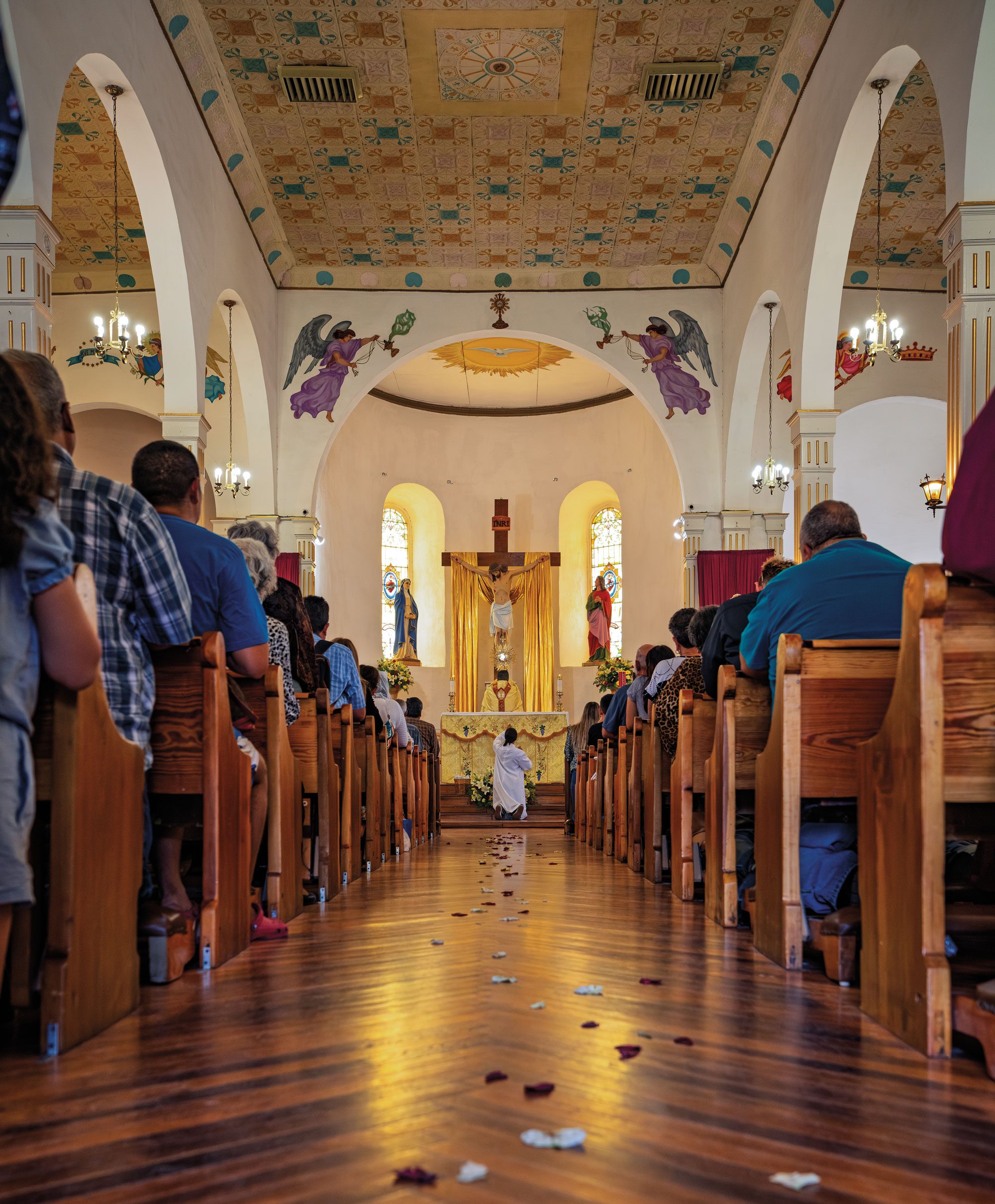 People sit in wooden pews leading up to an altar featuring a large crucifx. A man sits on his knees while praying in front of the altar inside a white church