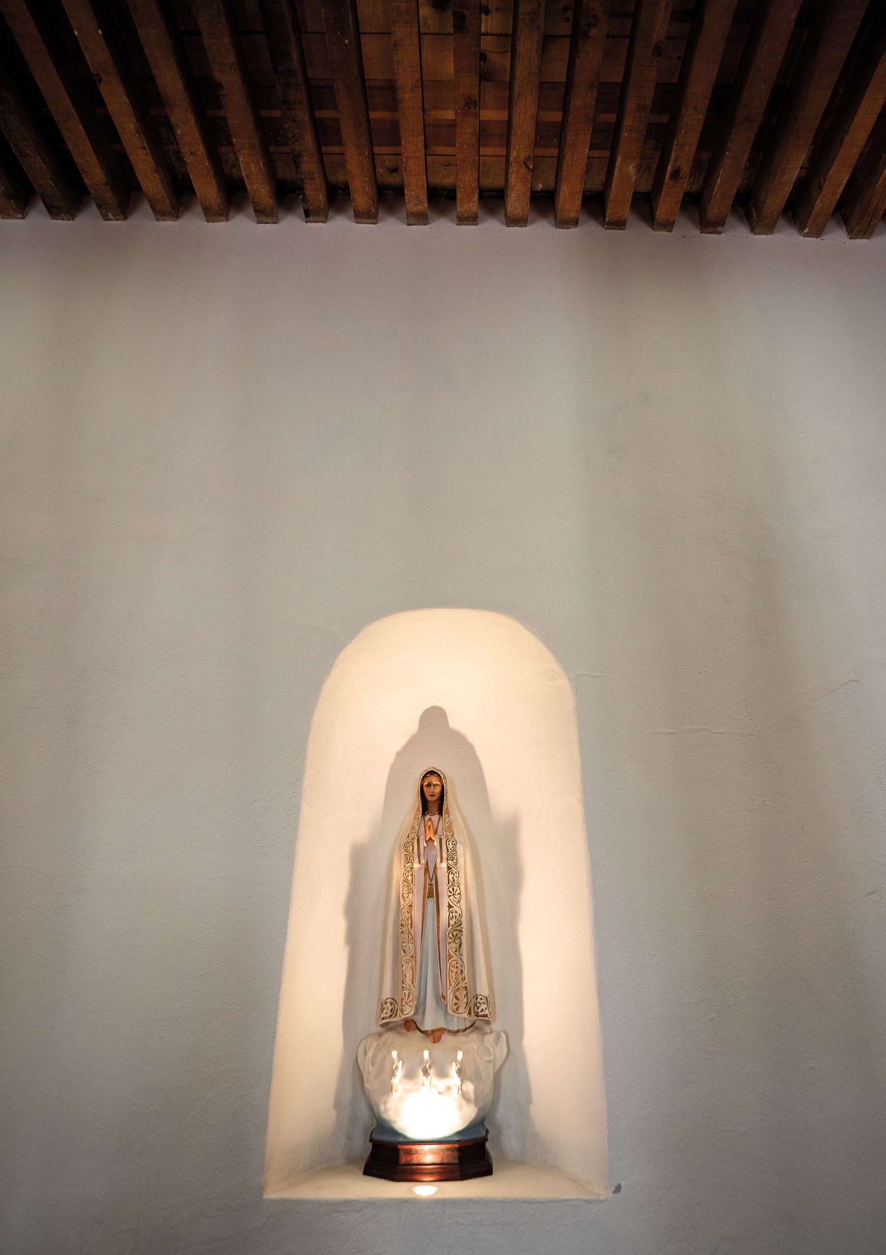 A small statue of the Virgin Mary dressed in a white rob sits illuminated inside a white adobe alcove.