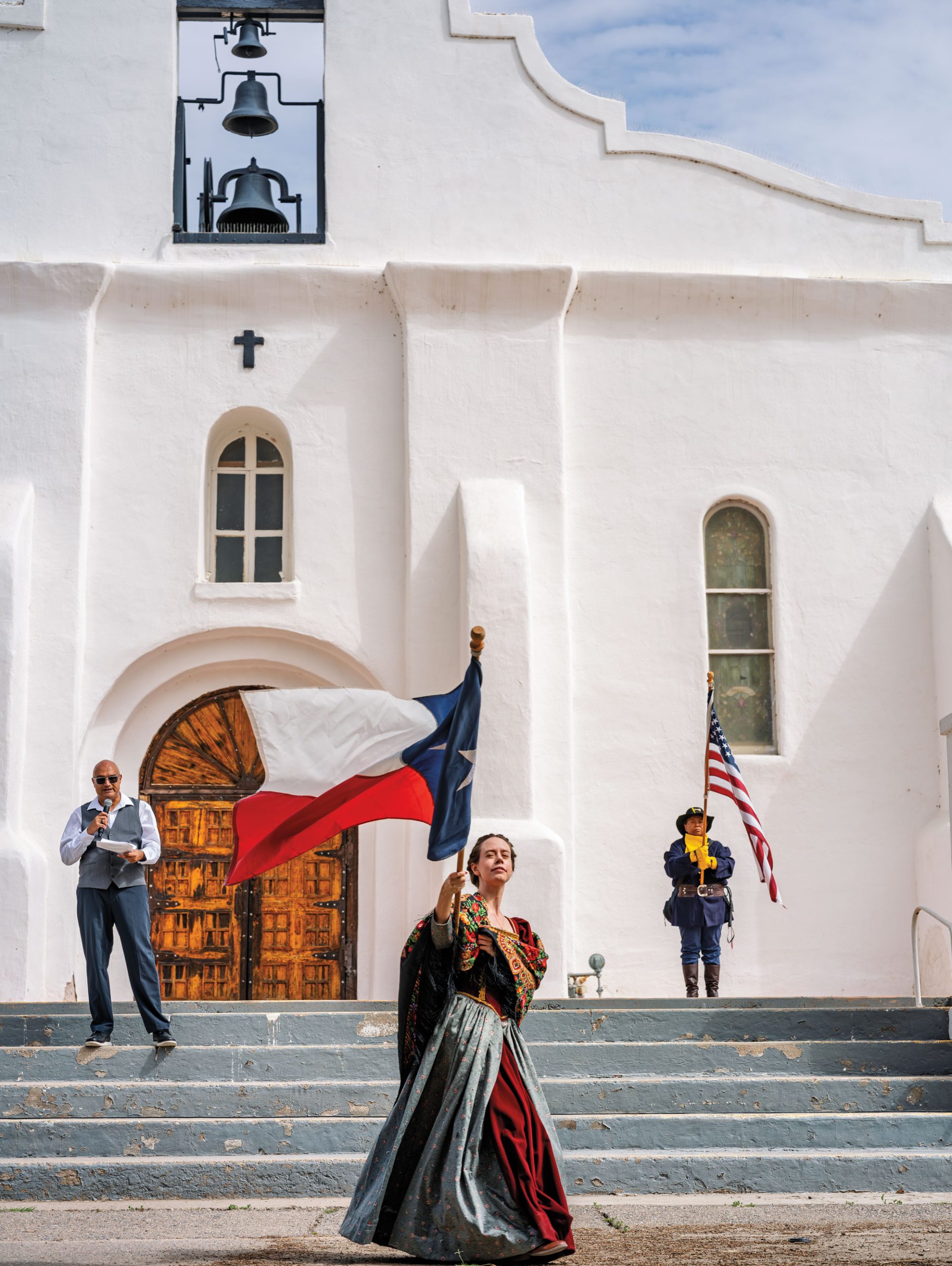 A woman in a red and blue dress stands in front of a white chapel waving a Texas flag. A man to the back right holds an American flag.
