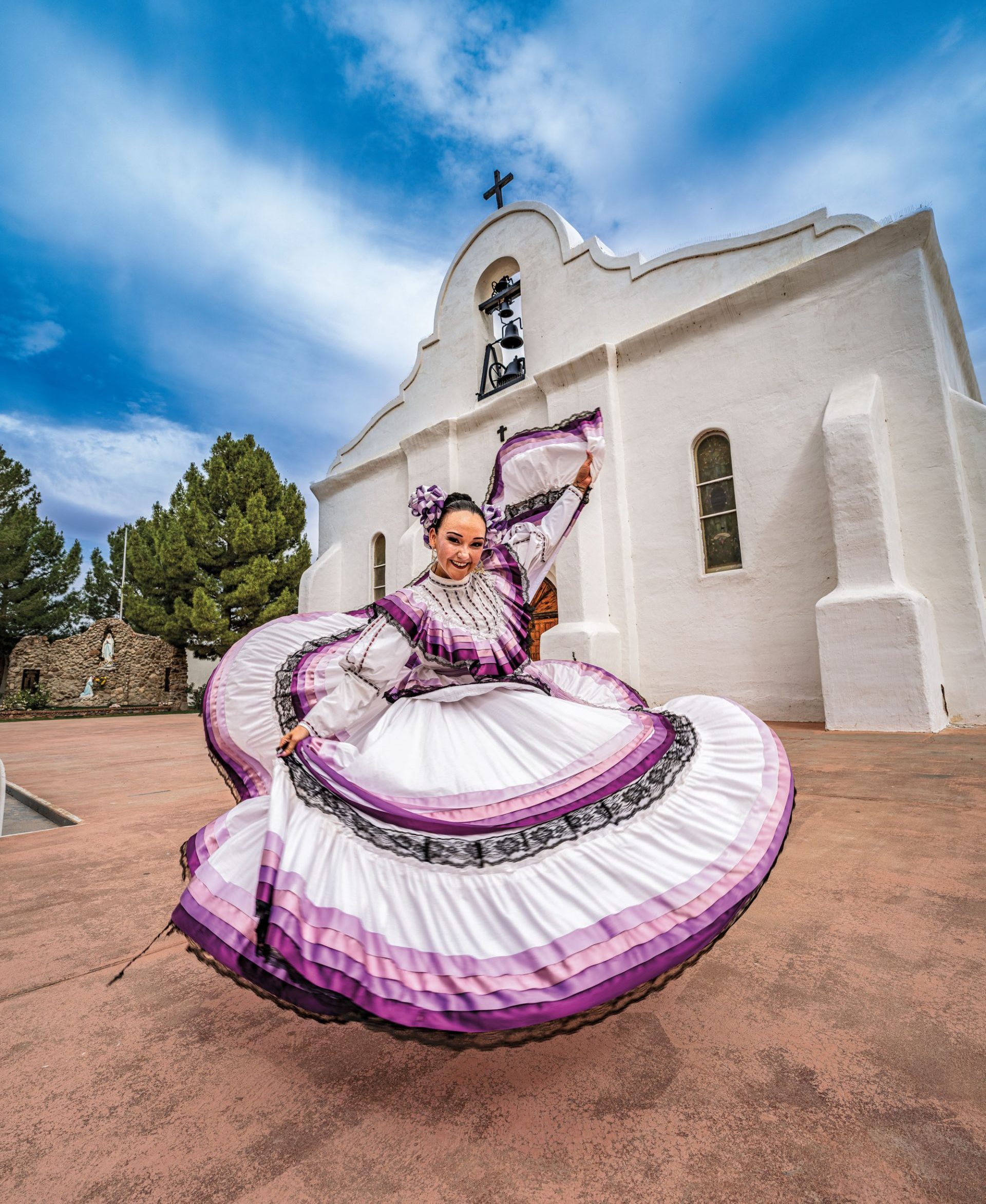 A woman in a purple and white traditional Mexican dance dress twirls her skirt and smiles in front of a white adobe chapel.