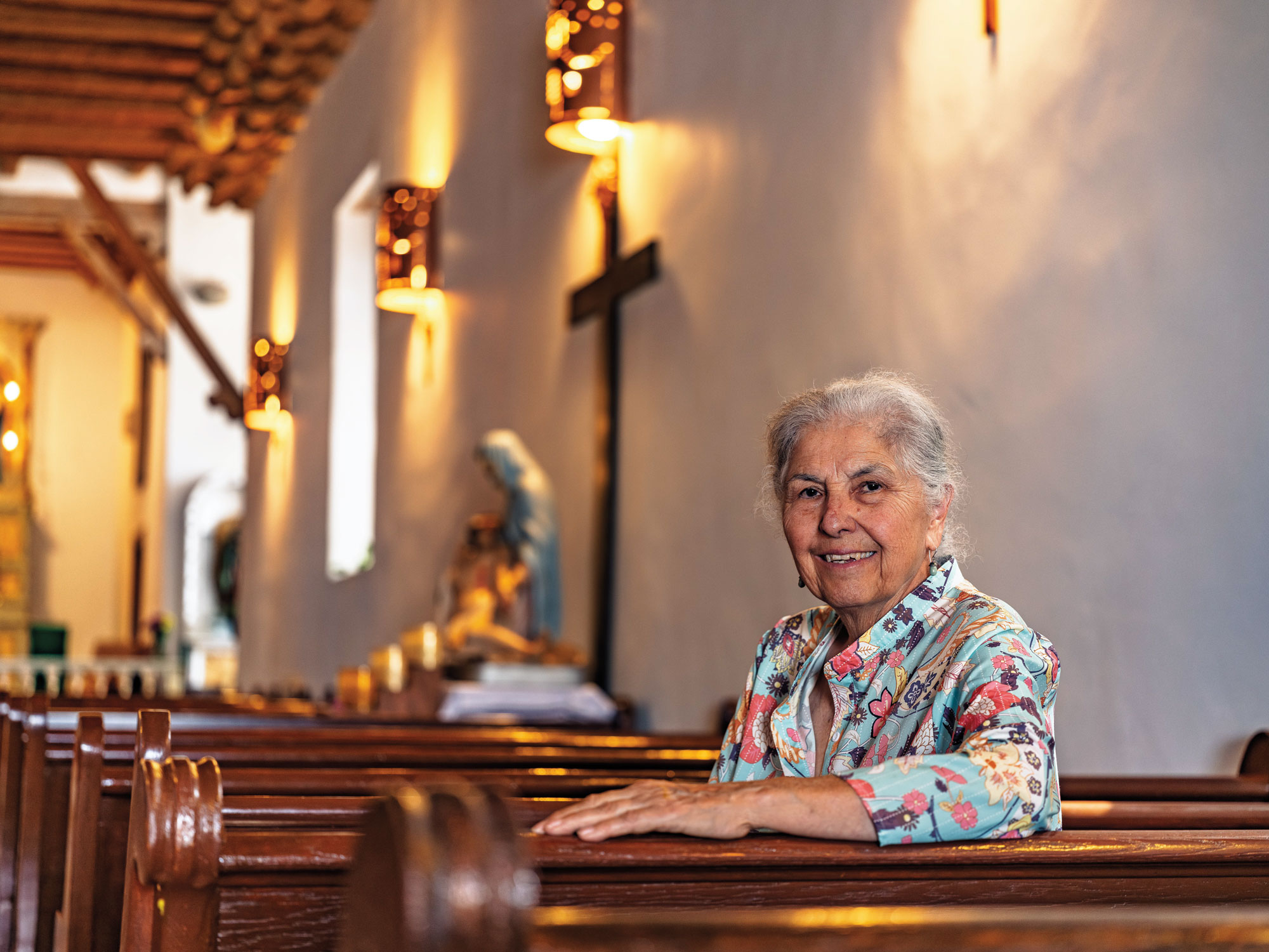 A woman with grey hair and a floral blouse smiles while sitting in a church pew