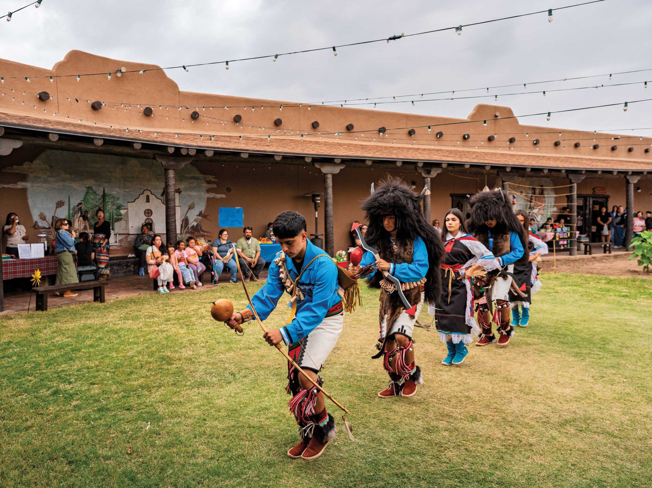 Five people in a row wearing colorful indigenous costumes that include belts, fringe worn around the legs, and headdresses