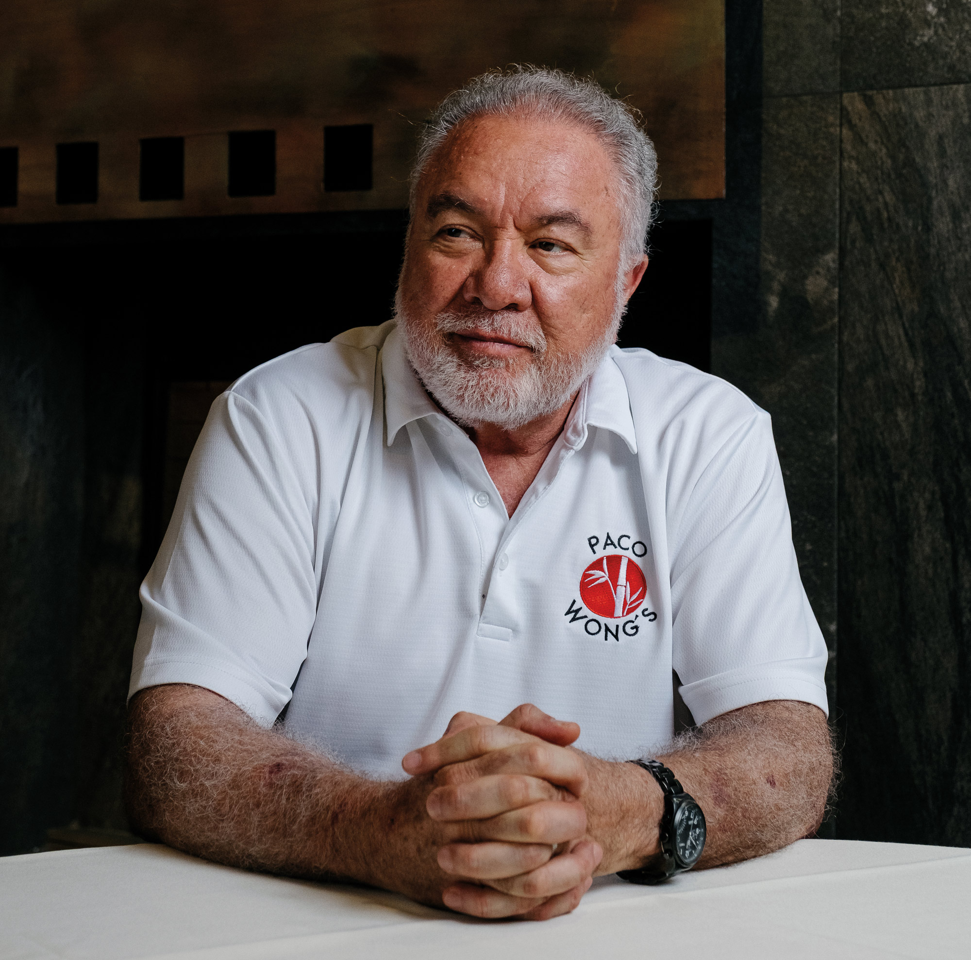A seated man with white hair and a white beard wearing a white polo shirt bearing the words Paco Wong's