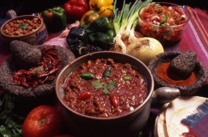 A bowl of chili with red and green peppers in it, surrounded by bowls of condiments like beans, diced tomatoes, ground chili powder, and dried red chilies.