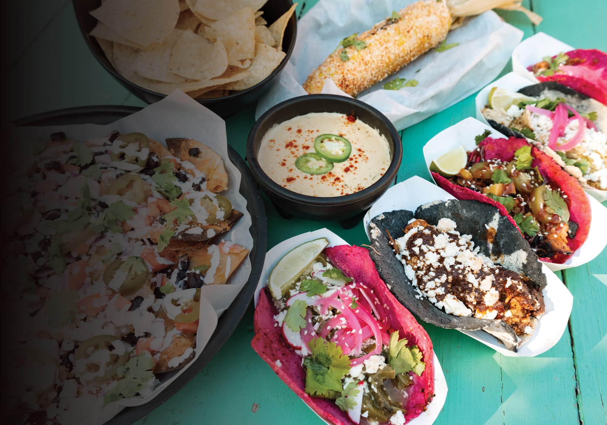 A teal picnic table filled with plates and baskets of food, including tacos with black and pink tortillas, nachos, queso, and tortilla chips.