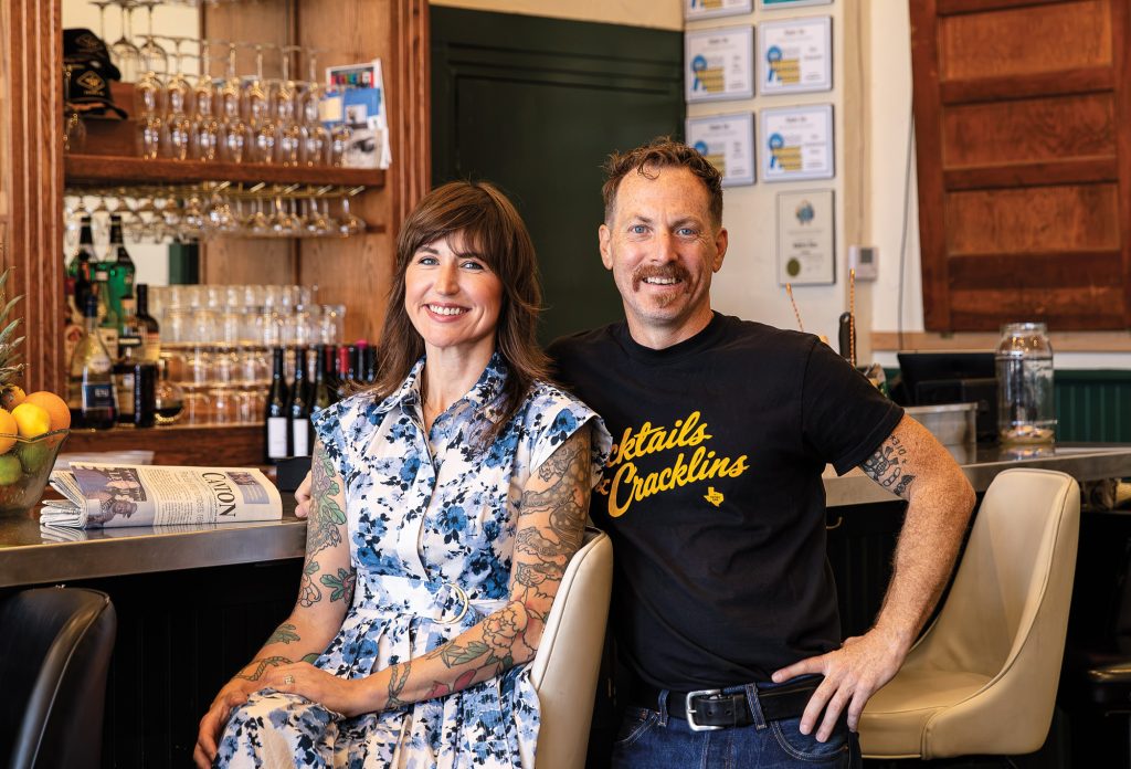 A brown-haired woman with tattoos on both arms next to a man in a T-shirt and blue jeans in front of a bar