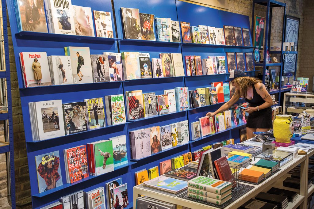 A person in a blue hat and black dress selecting a magazine from a large blue wall of magazines
